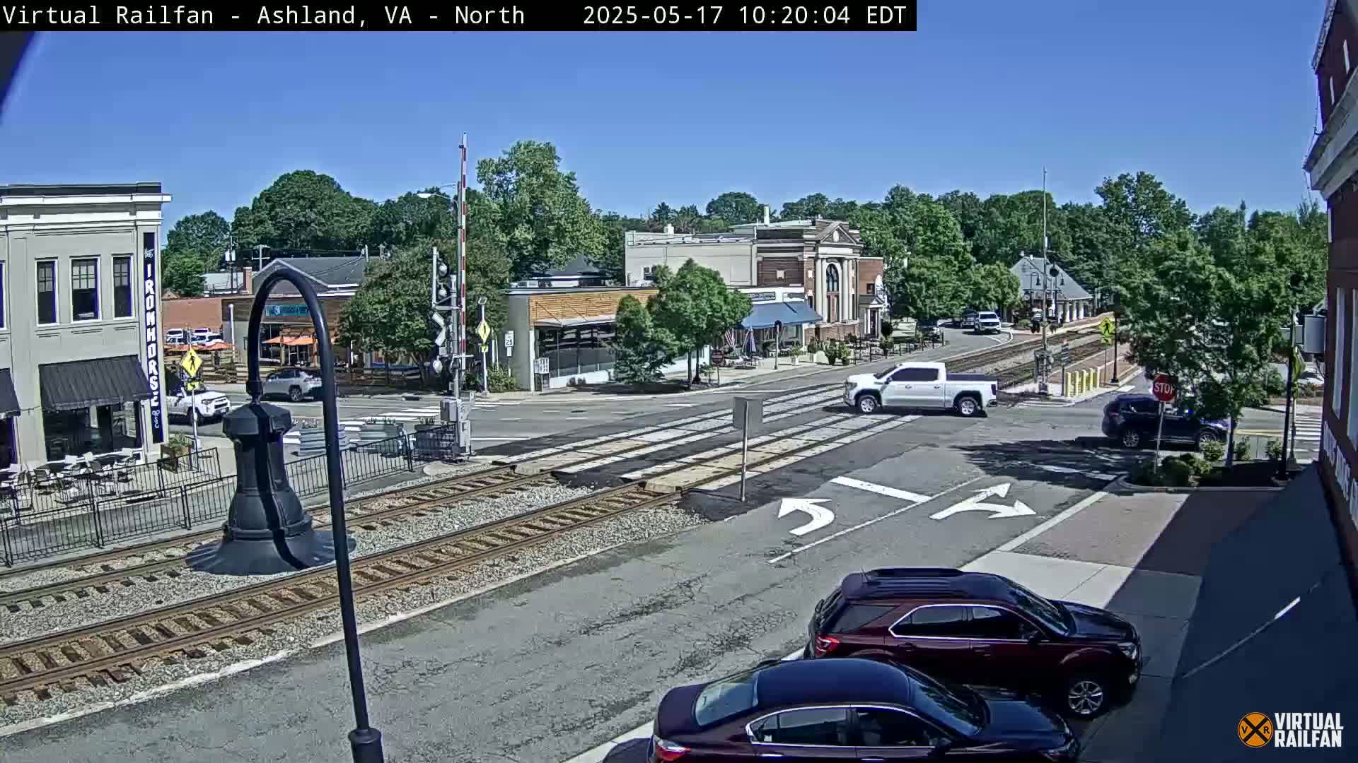 A sunny day shows a street intersection with train tracks, several cars, and buildings with outdoor seating.