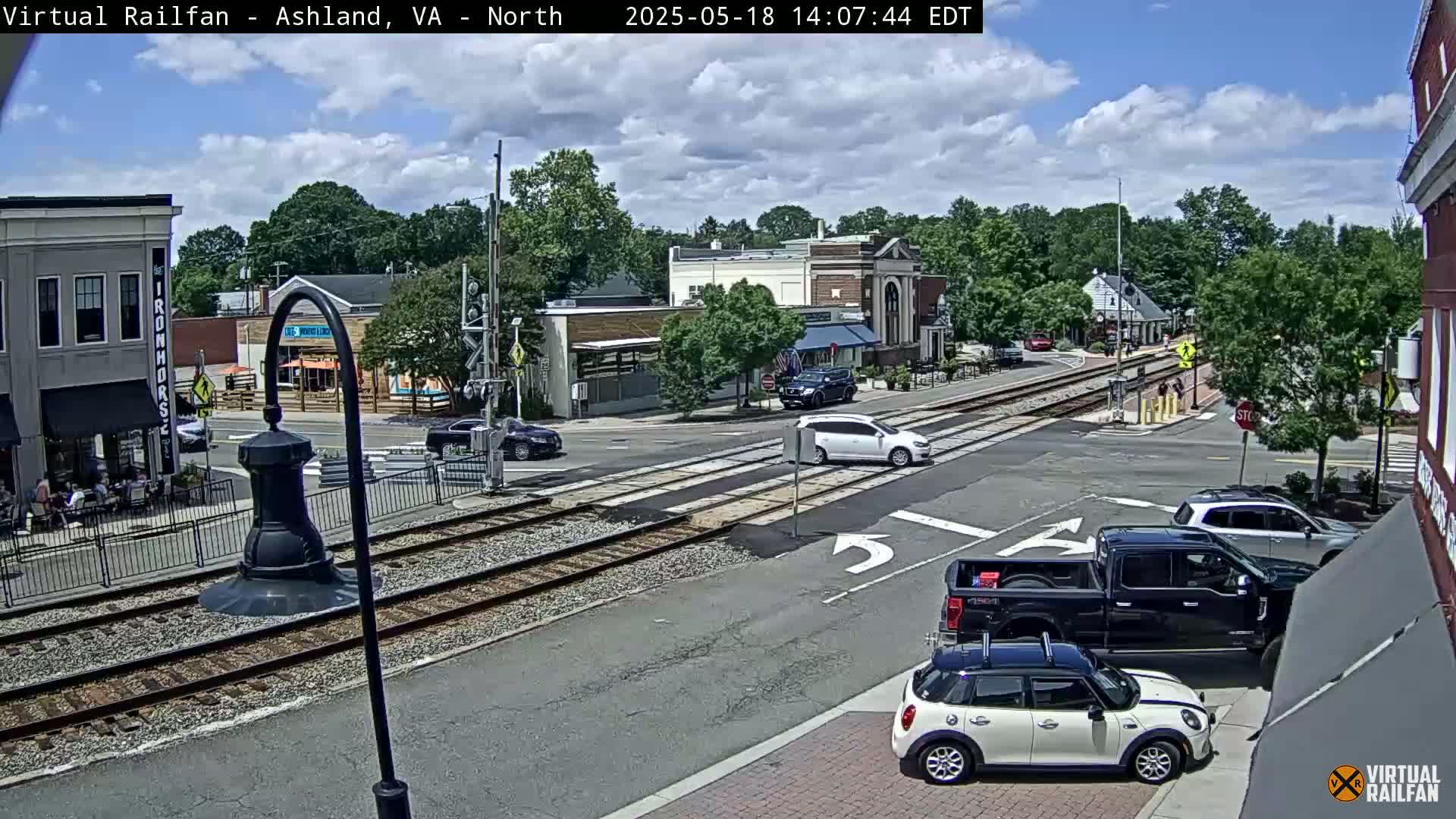 A partly cloudy day shows a town square with railroad tracks intersecting a street, several cars at a crossroads, and buildings lining the street.