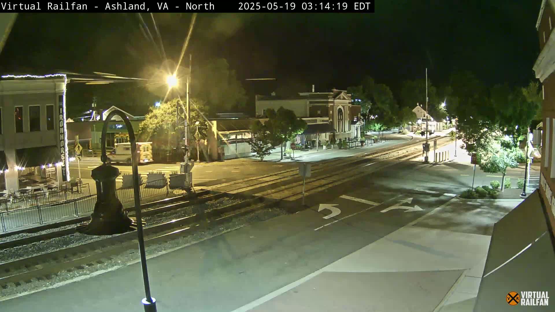 A nighttime view of a town's street intersection with train tracks, featuring buildings with streetlights illuminating the scene.