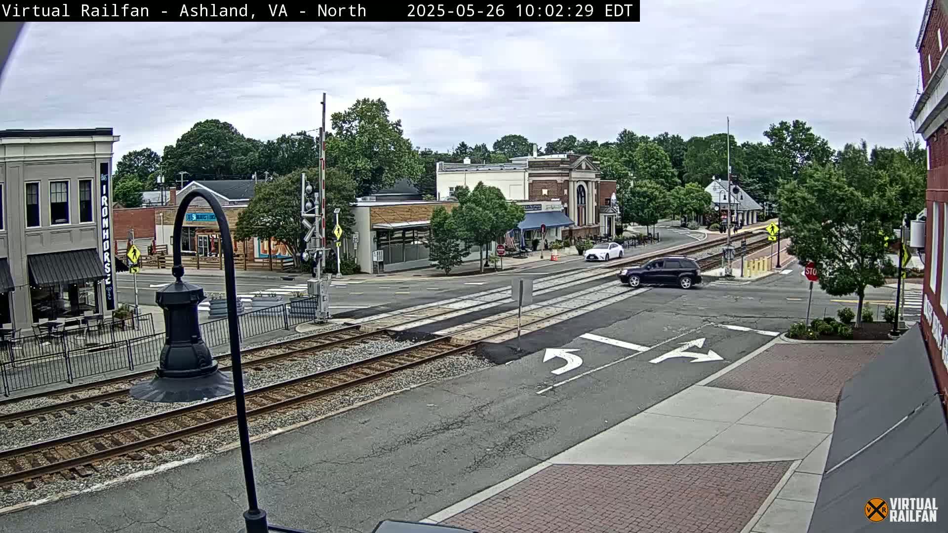 A partly cloudy day shows a view of a town's intersection with railroad tracks, featuring several buildings and a few cars.