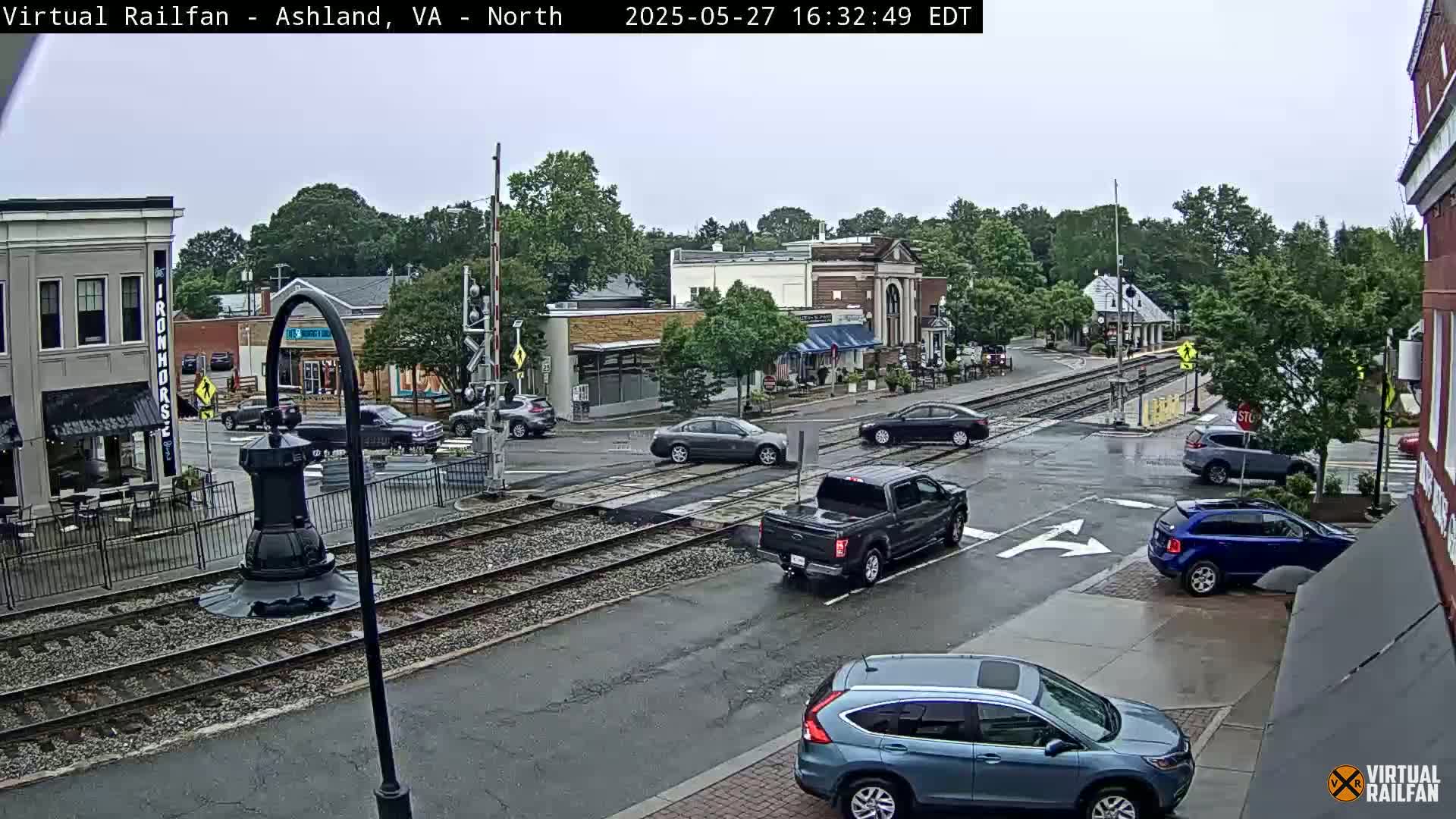 A rain-slicked town square with cars at an intersection near train tracks, surrounded by low buildings and trees.