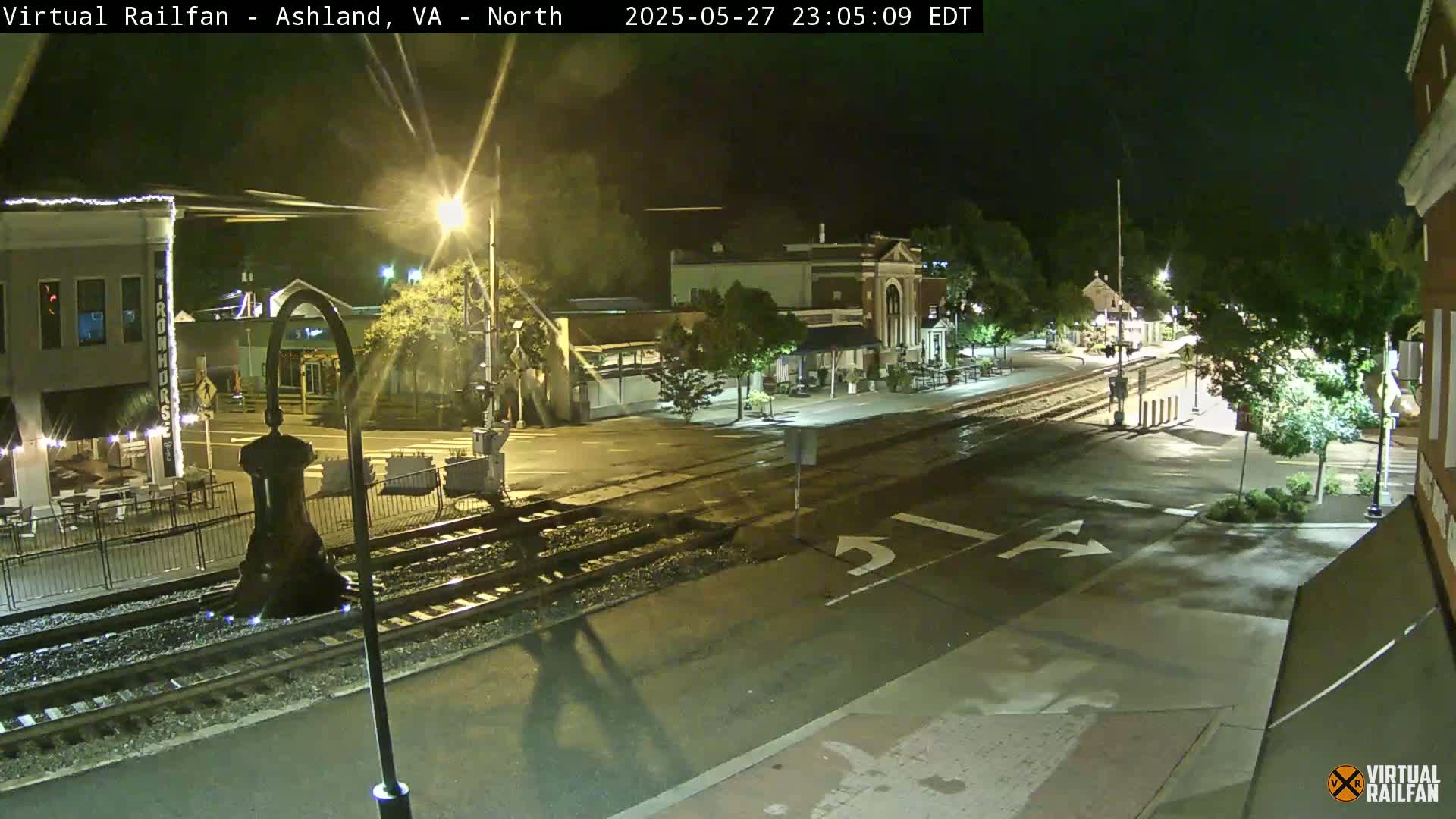 A nighttime view of a town's street intersection with train tracks, featuring buildings and streetlights under partly cloudy skies.