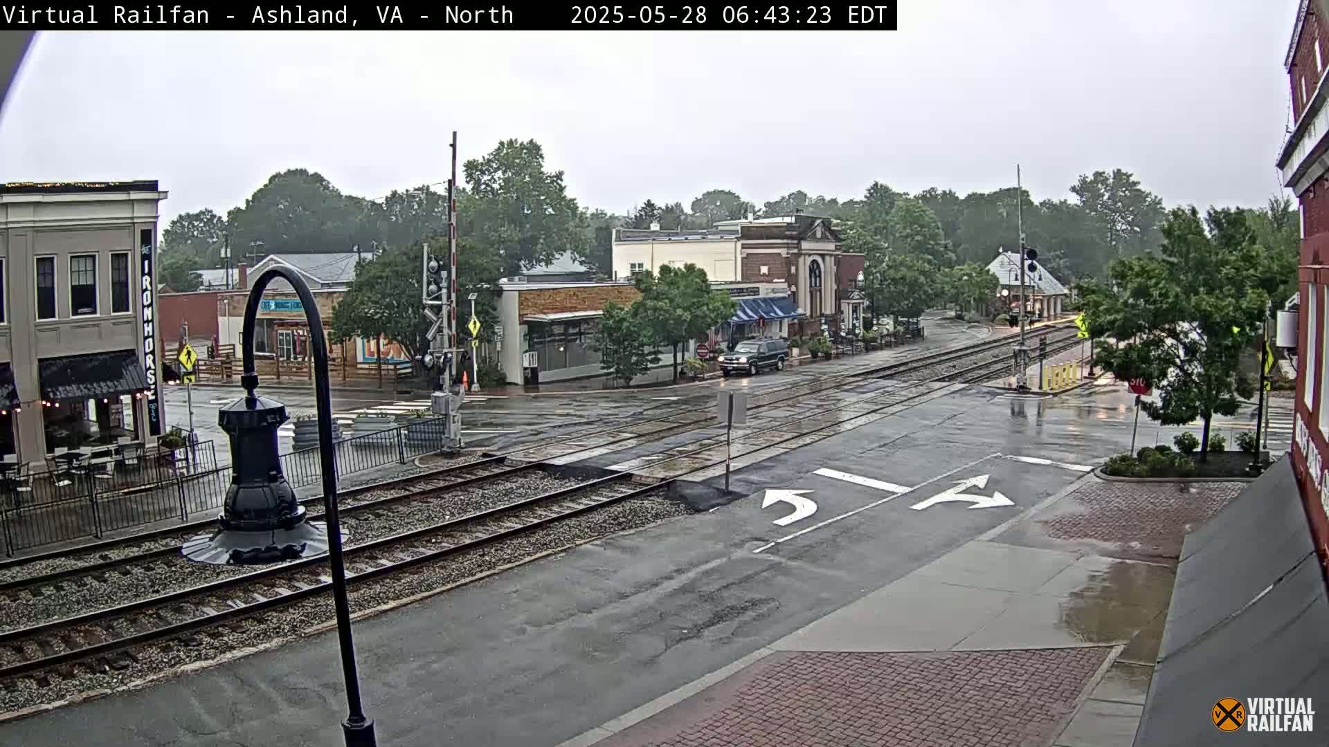 A wet town square with railroad tracks intersecting a street, surrounded by buildings and trees under an overcast sky.
