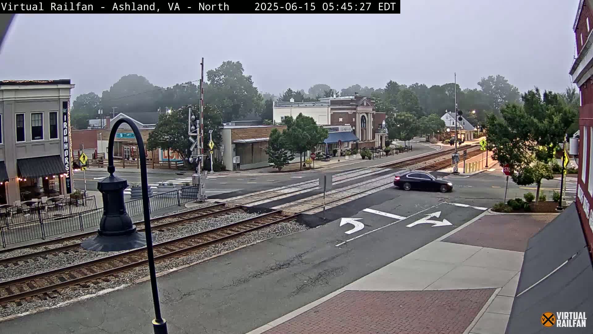 A foggy morning scene shows a street intersection with train tracks, several buildings, and a single dark-colored car.