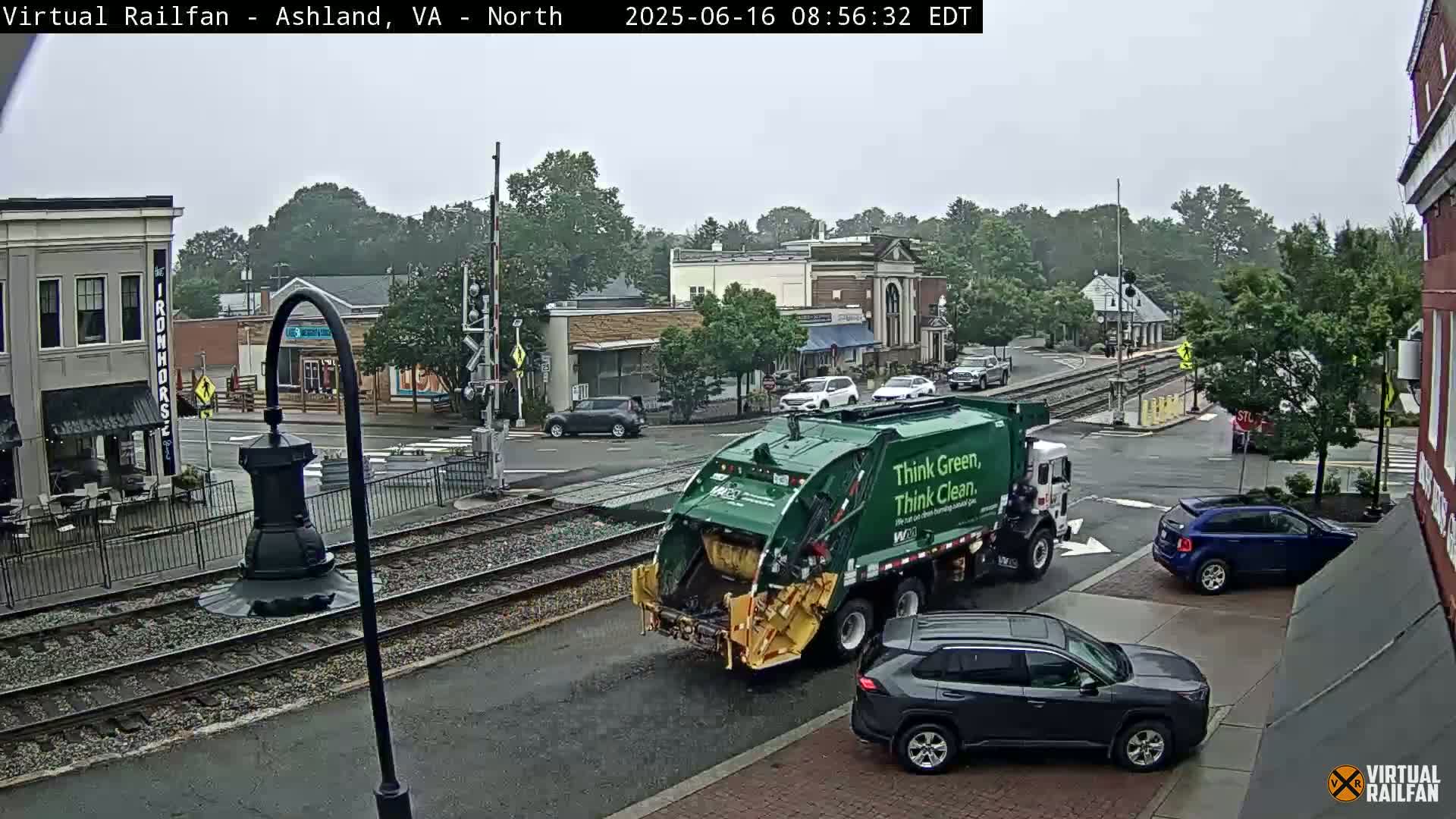 A green garbage truck and several cars are on a street in a town, under an overcast sky.