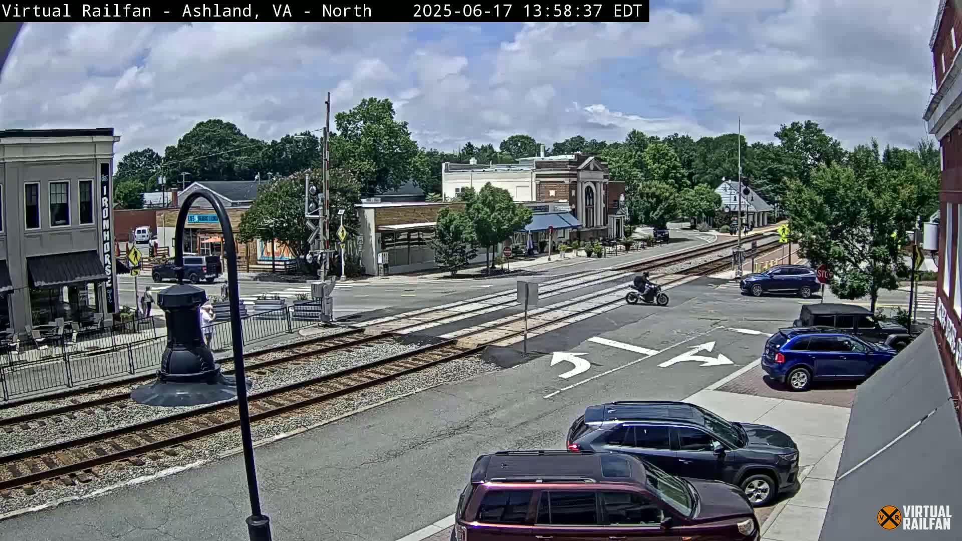 A partly cloudy day shows a view of a town's intersection with railroad tracks, cars parked and driving, and a motorcyclist; some pedestrians are also visible.