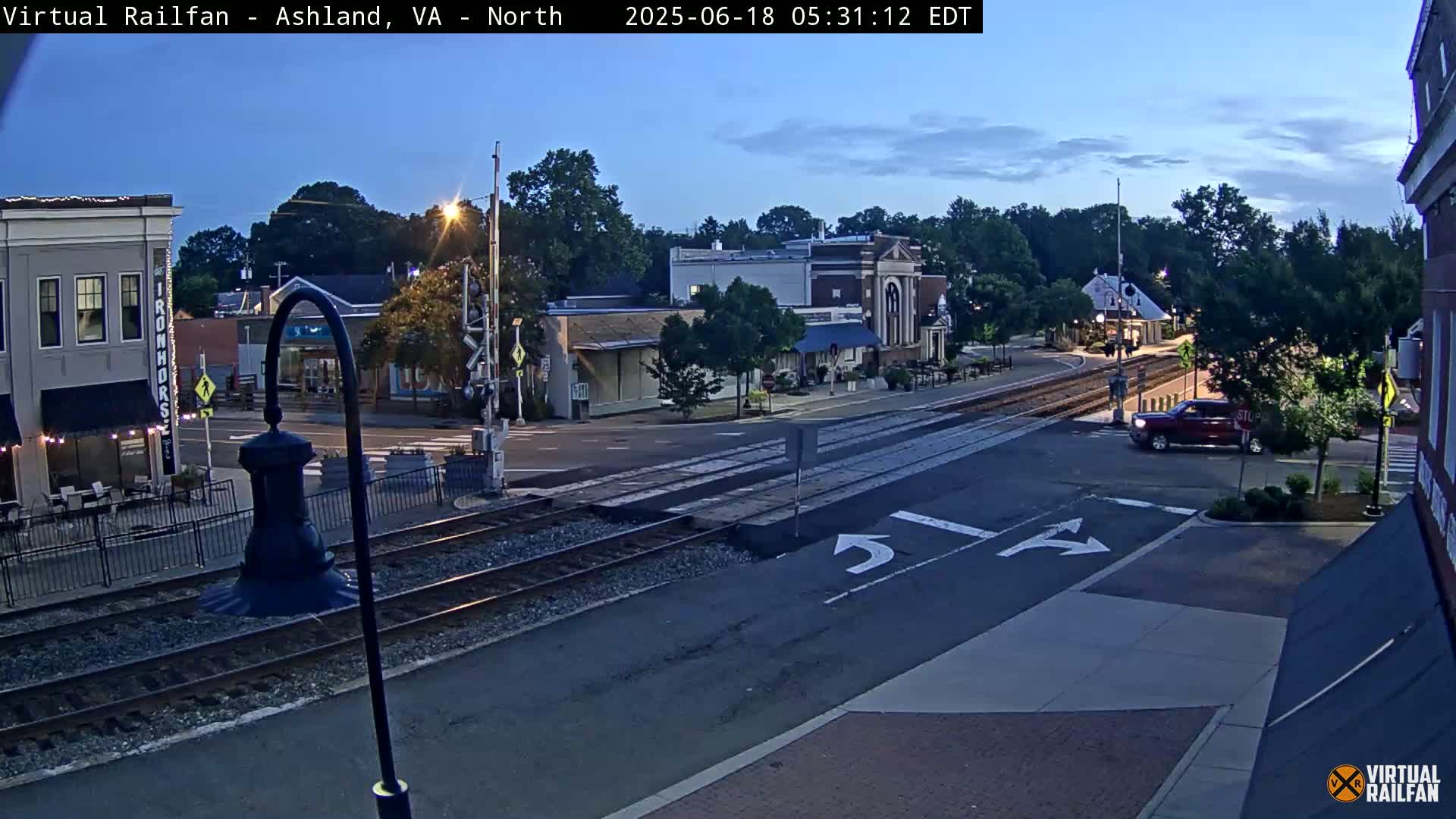 A twilight view of a town's street intersection with railroad tracks, featuring several buildings, trees, and a red truck.