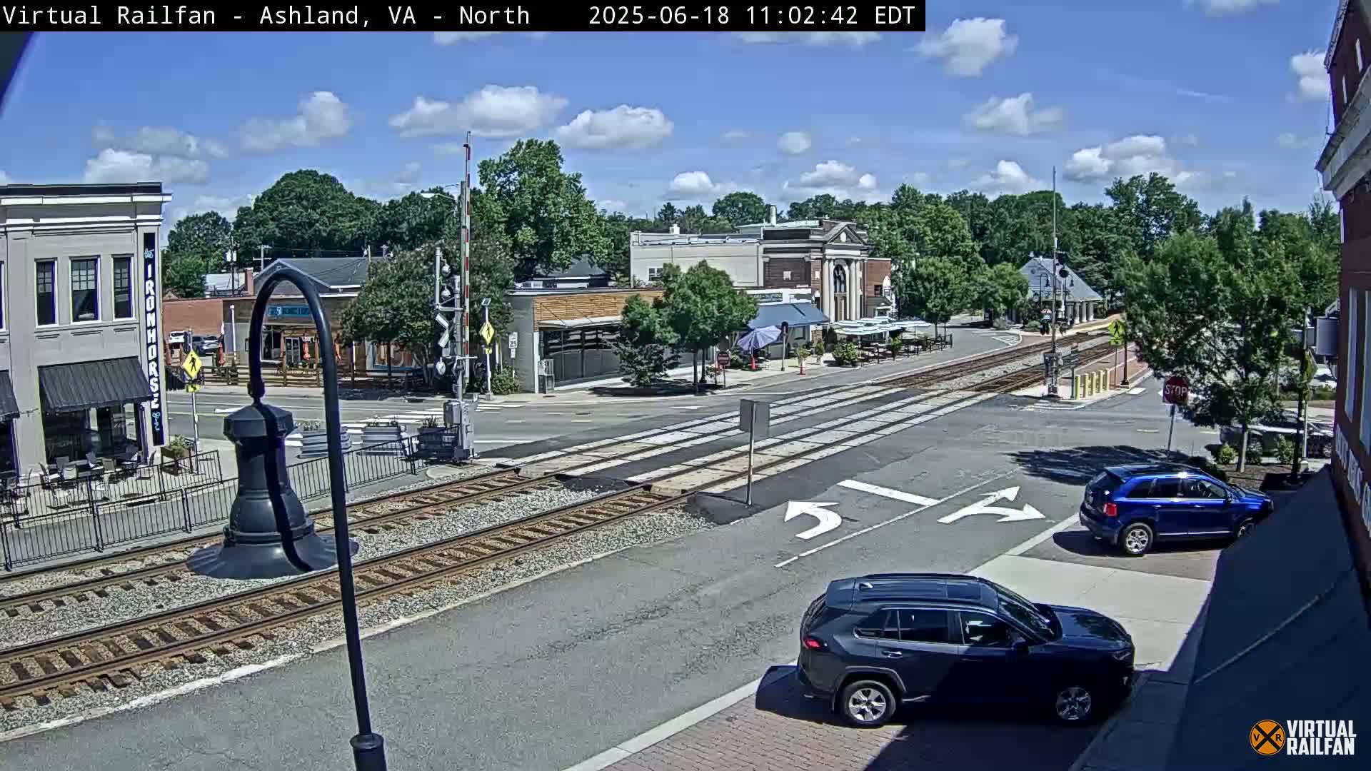 A sunny day view of a town's street intersection with railroad tracks, featuring several buildings and a couple of cars, one blue and one dark-colored.