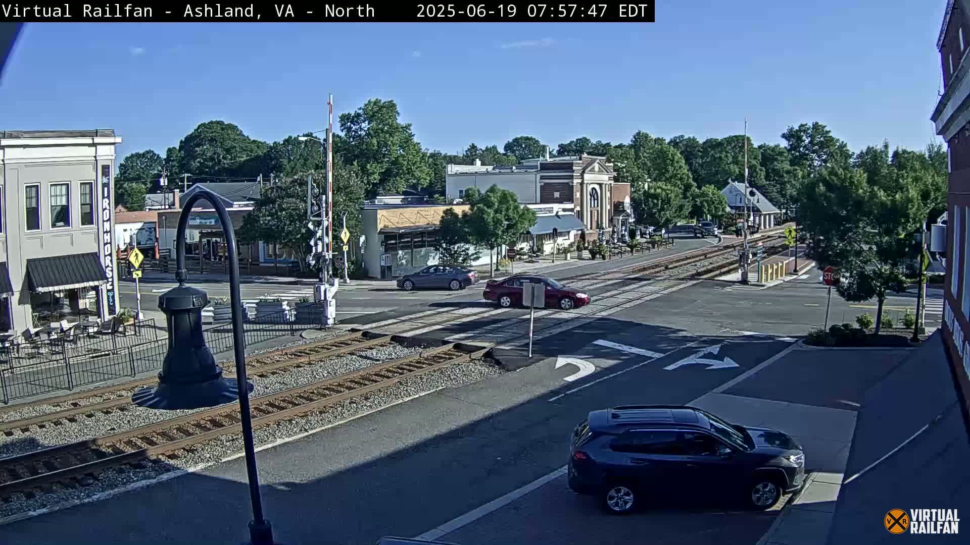 A sunny day shows a railroad crossing in a town with several cars, buildings, and trees.