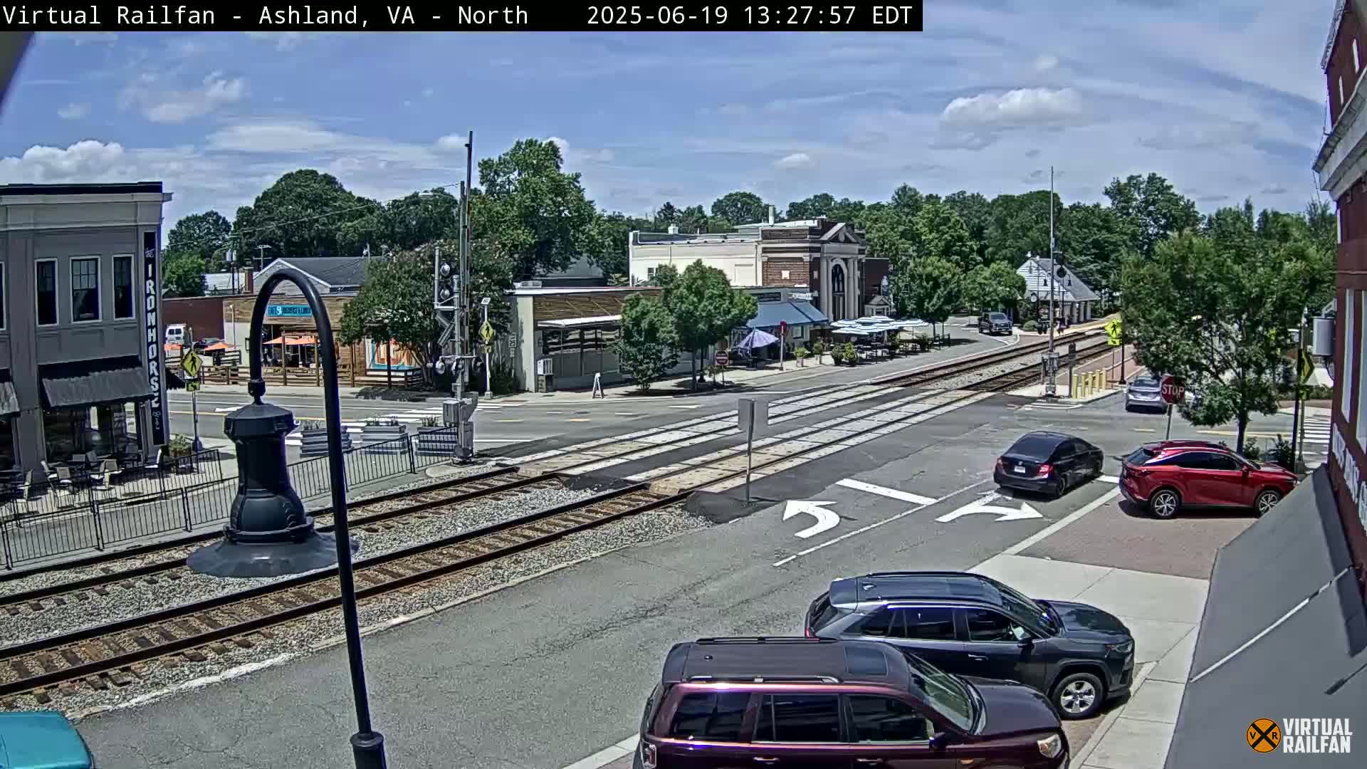 A sunny day shows a street intersection with railroad tracks, several cars, and buildings with outdoor seating areas.