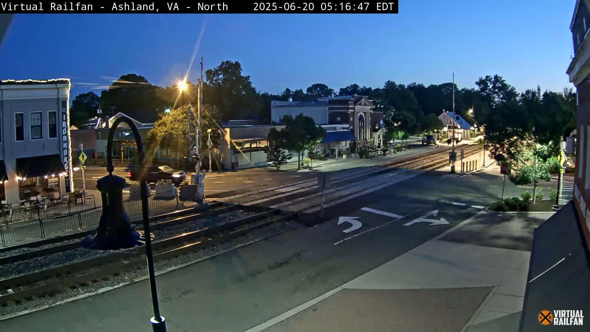 A twilight view of a town's street intersection with train tracks, featuring several buildings and streetlights under a clear sky.