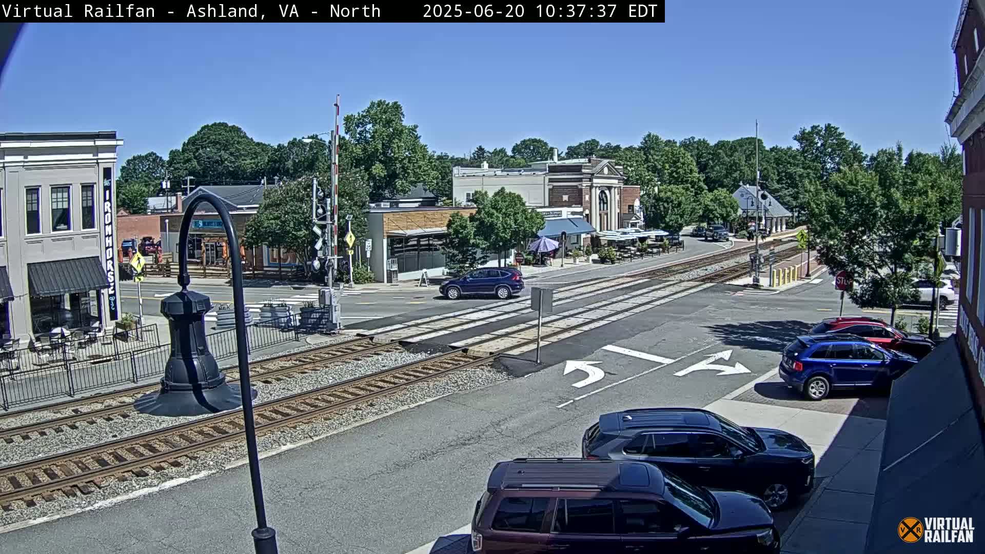 A sunny day shows a street scene with railroad tracks crossing a road in front of several buildings and cars parked along the street and several cars moving through the intersection.