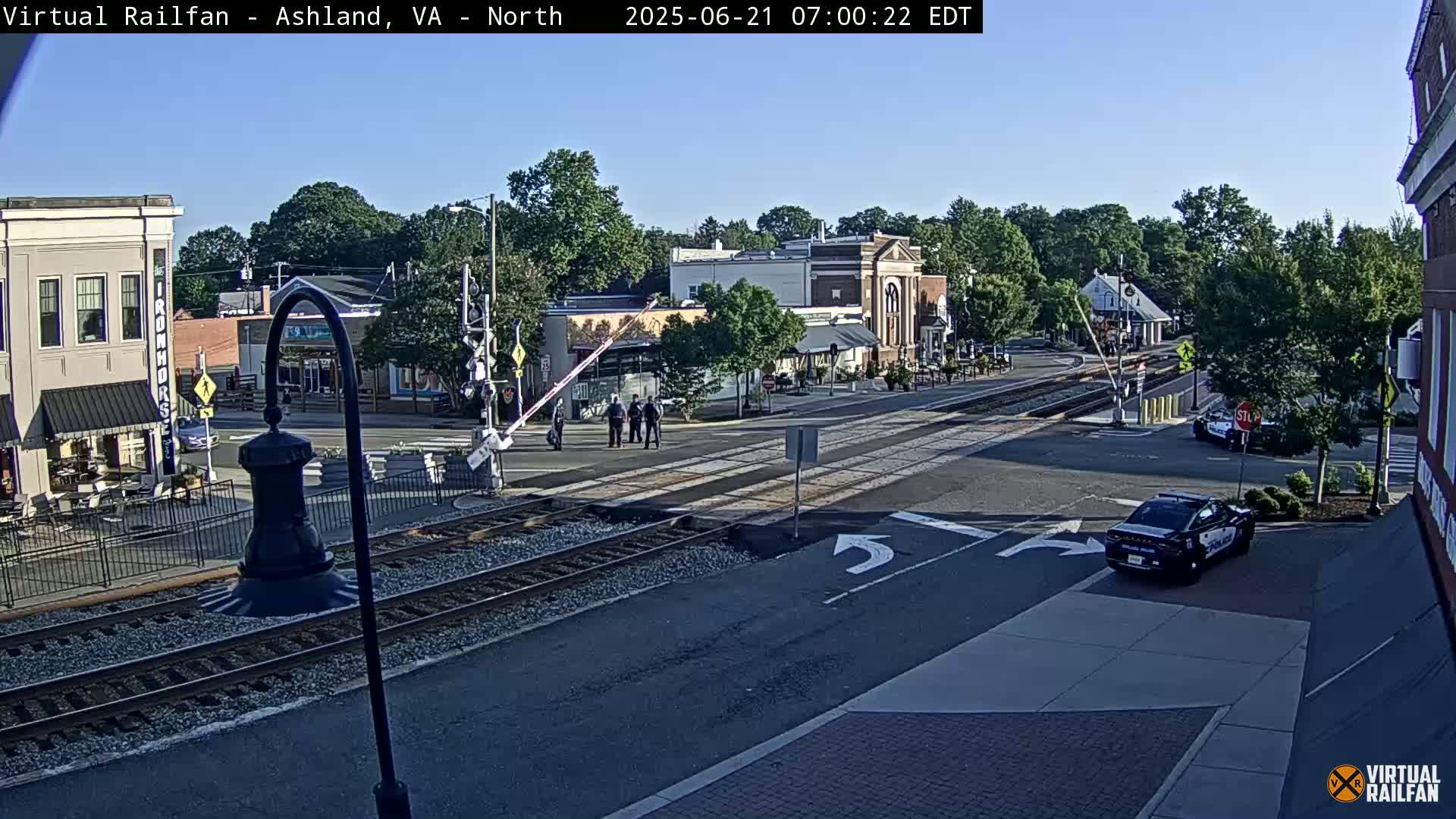 A sunny day view of a town's railroad crossing with several buildings, a few pedestrians, and police cars present.
