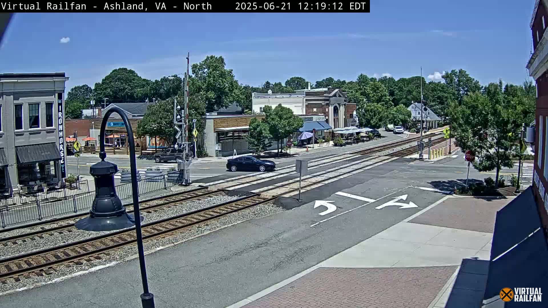 A sunny day shows a view of a town's intersection with train tracks, buildings, and cars, under a clear blue sky.
