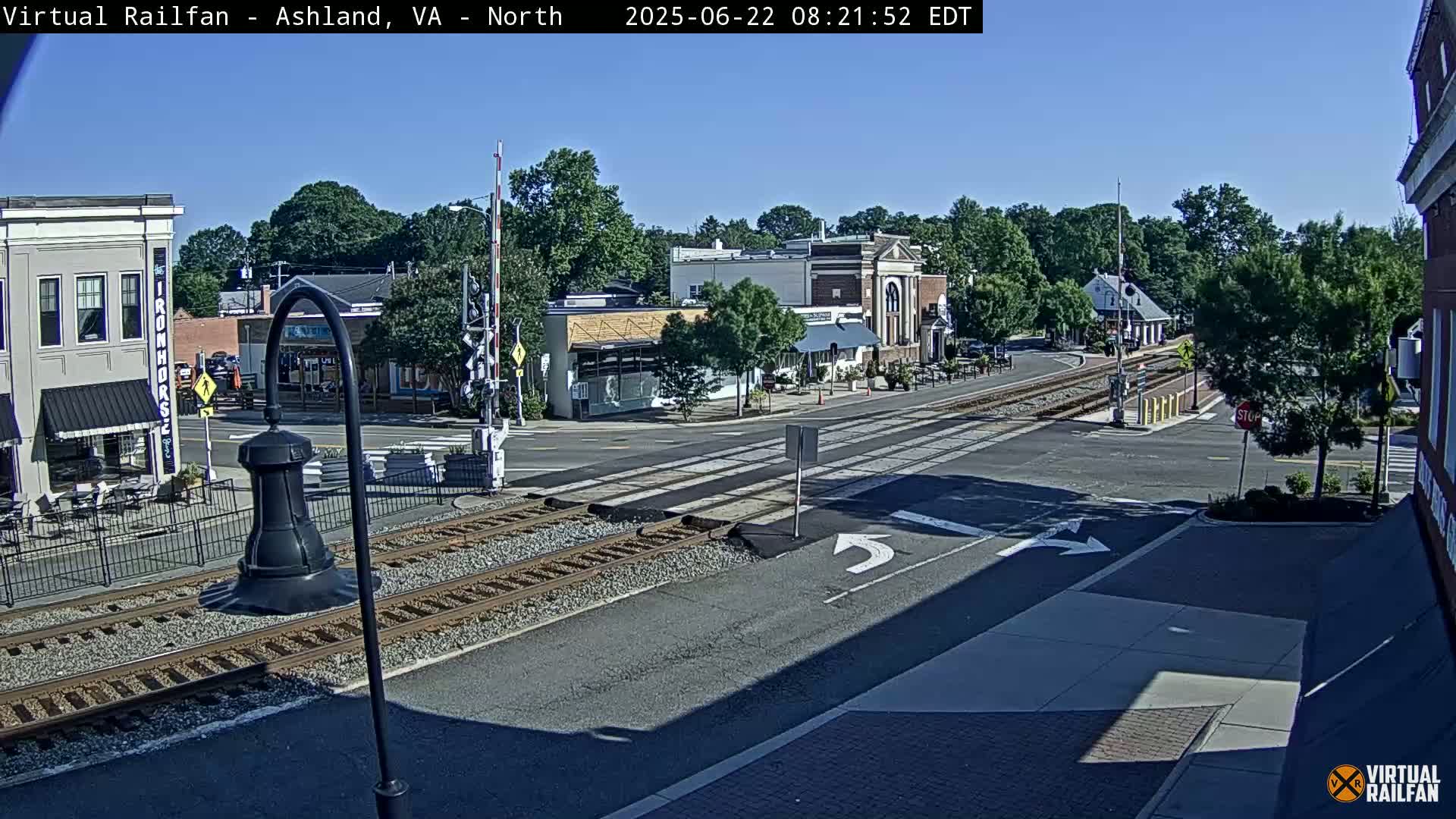 A sunny day reveals a railroad crossing in a small town, where a street intersects train tracks near several buildings and lush green trees.
