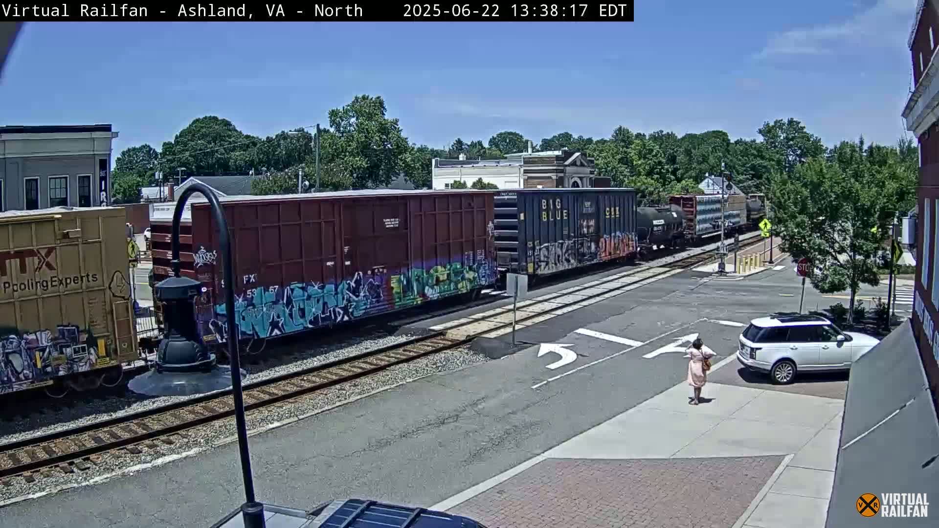 A freight train with graffiti-covered cars passes through a town intersection on a sunny day, as a person walks across the street near a white SUV.