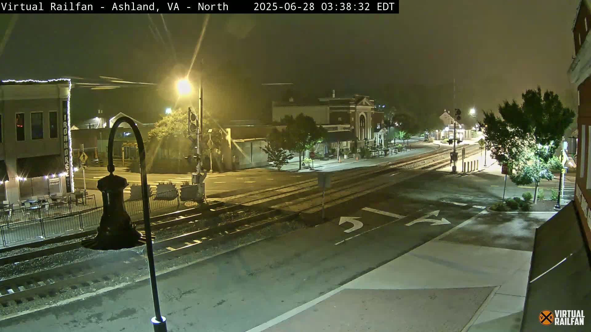 A nighttime view of a town's street intersection with railroad tracks, under hazy conditions, showing buildings, streetlights, and trees.