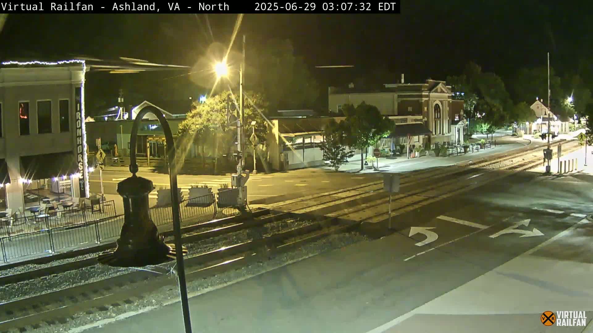 A nighttime view of a town's street intersection with train tracks, showing several buildings and streetlights.