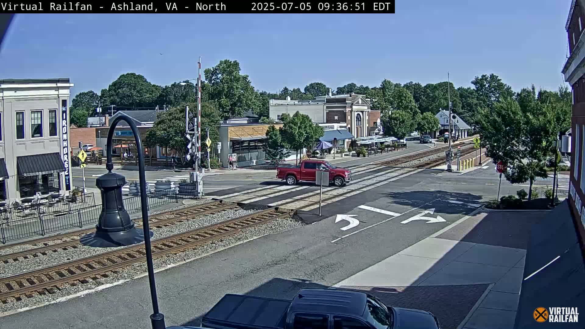 A sunny day shows a red pickup truck crossing railroad tracks at an intersection in a town with several buildings, some with outdoor seating.