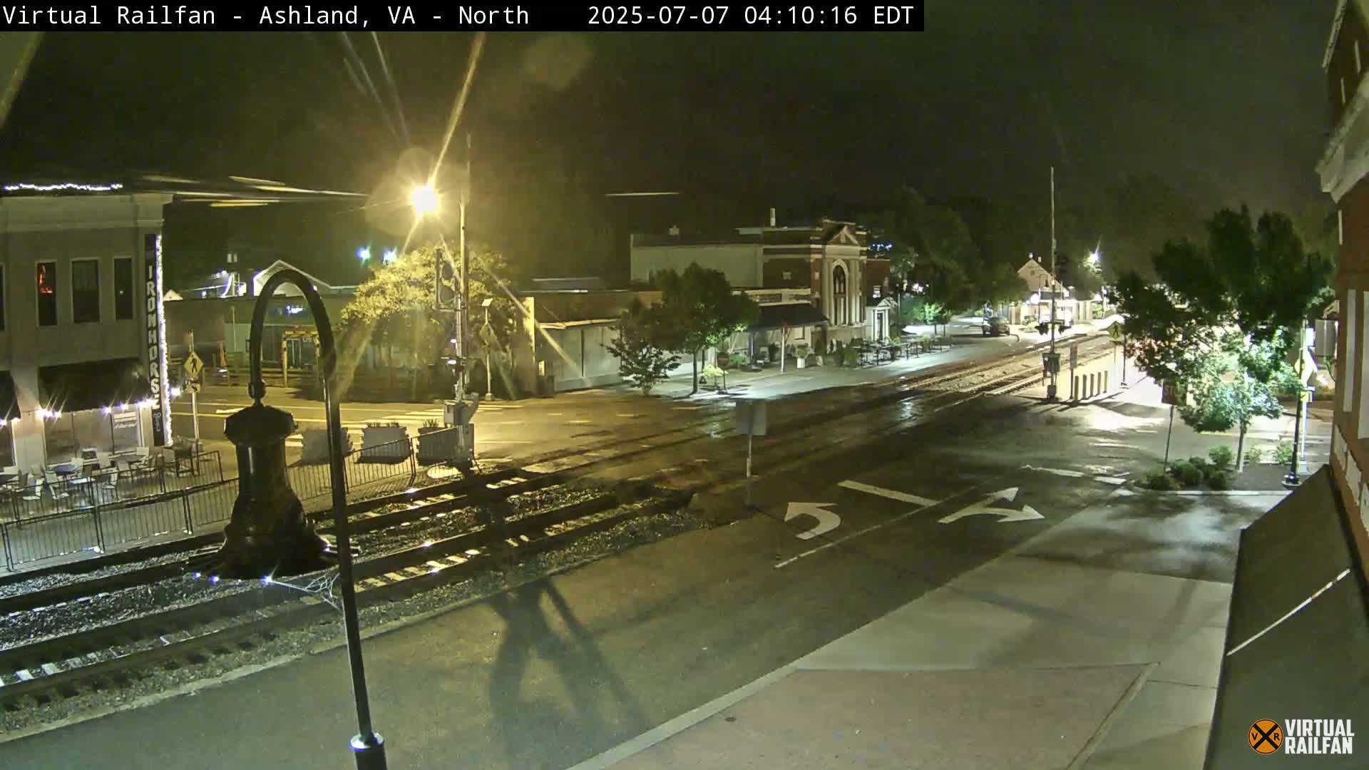 A rain-slicked, nighttime street scene shows a railroad crossing in front of several low-rise buildings and trees.