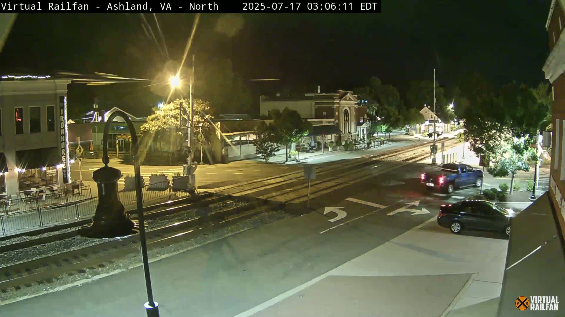 A nighttime street scene shows a railroad crossing in a small town with a few cars and buildings illuminated by streetlights.