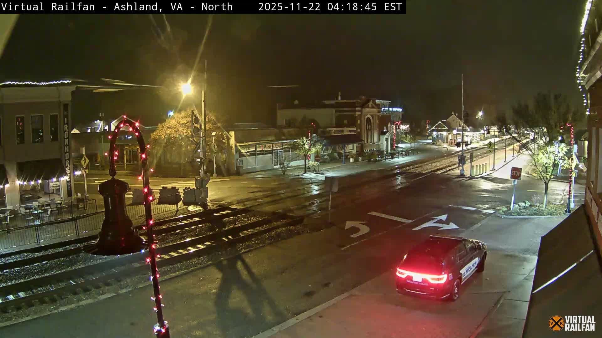 A police car with illuminated taillights drives on a wet street at night, passing railroad tracks and buildings adorned with holiday lights under the glow of streetlights.