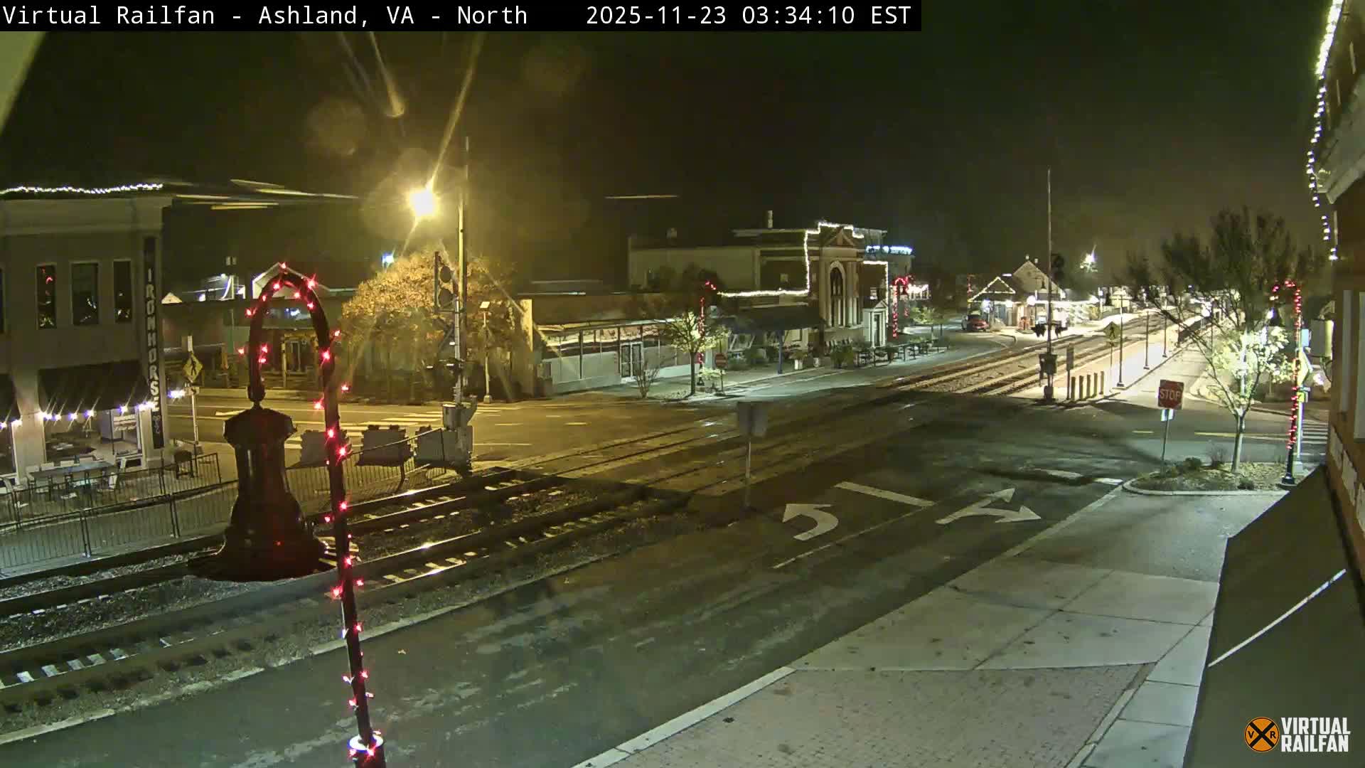 The image displays a damp, clear night scene of a town street with railroad tracks crossing an intersection, flanked by buildings decorated with festive string lights and illuminated by streetlights.
