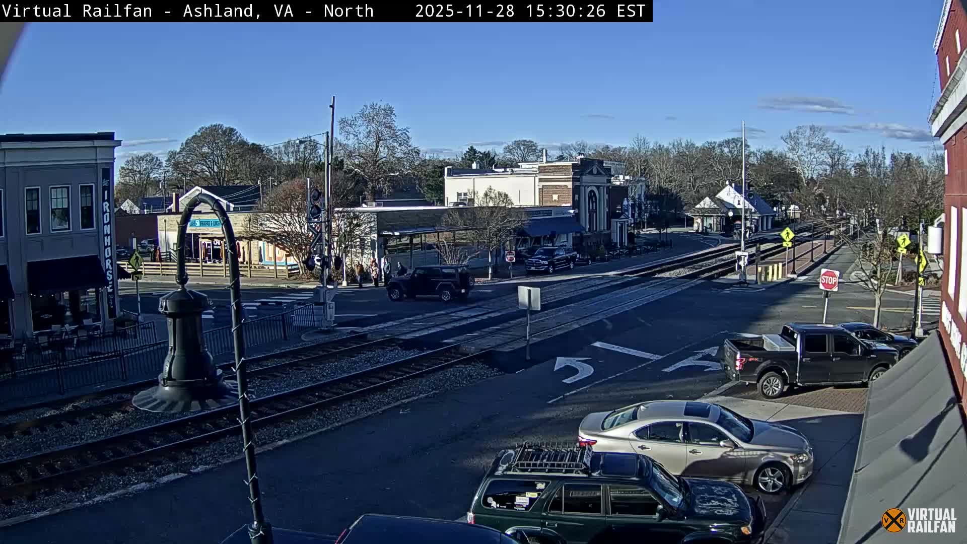 A sunny, clear day illuminates a town street with a double set of train tracks intersecting a road bustling with several parked and moving cars, bordered by buildings and mostly bare trees.