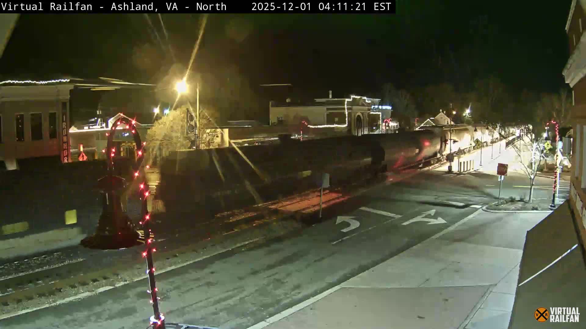 A long train is passing through a brightly lit downtown area adorned with holiday decorations at night, under clear skies.