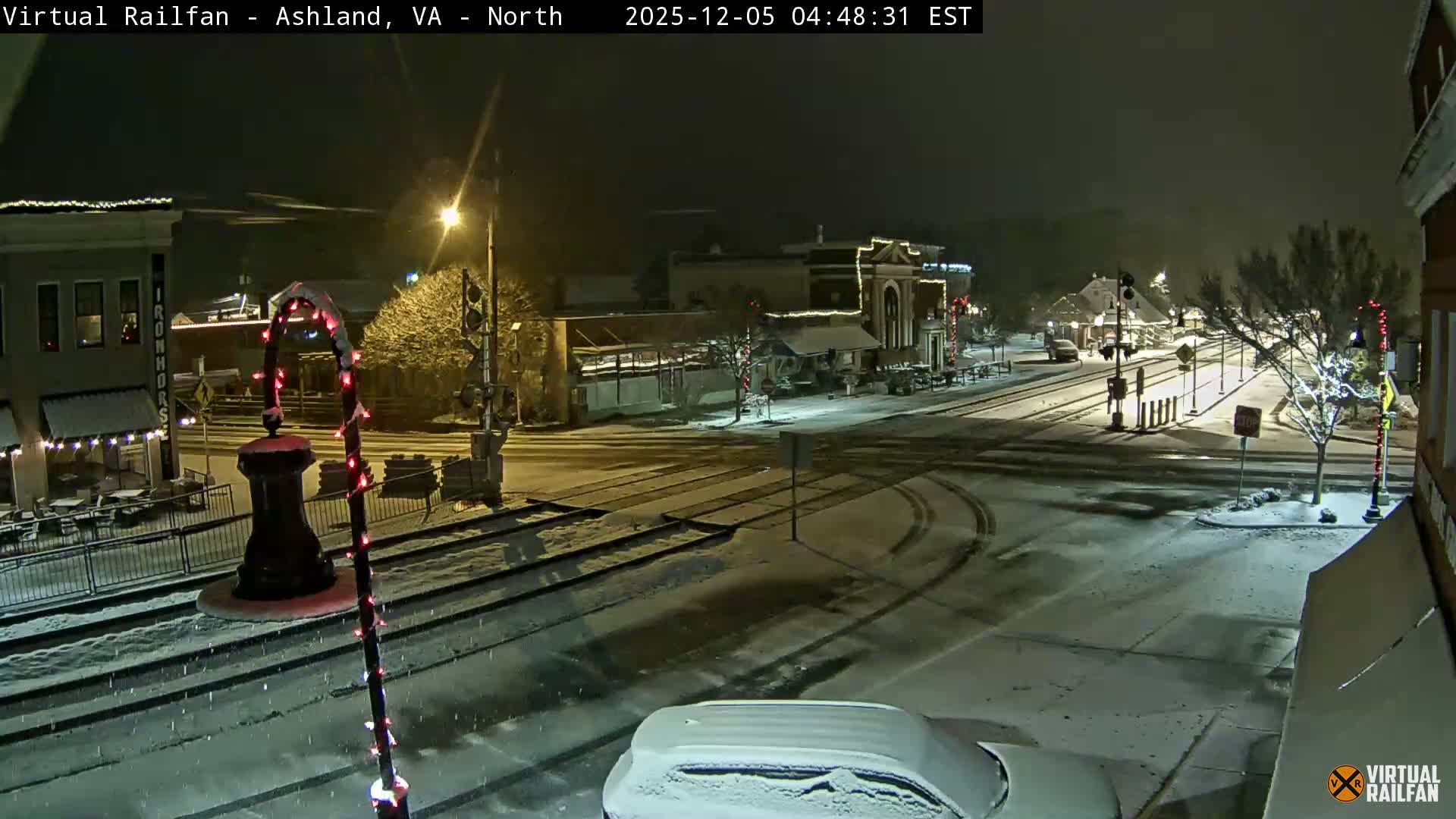 A wintry night scene shows a snow-covered town street with railroad tracks, buildings adorned with festive lights, and a vehicle, all illuminated under a blanket of fresh snow.