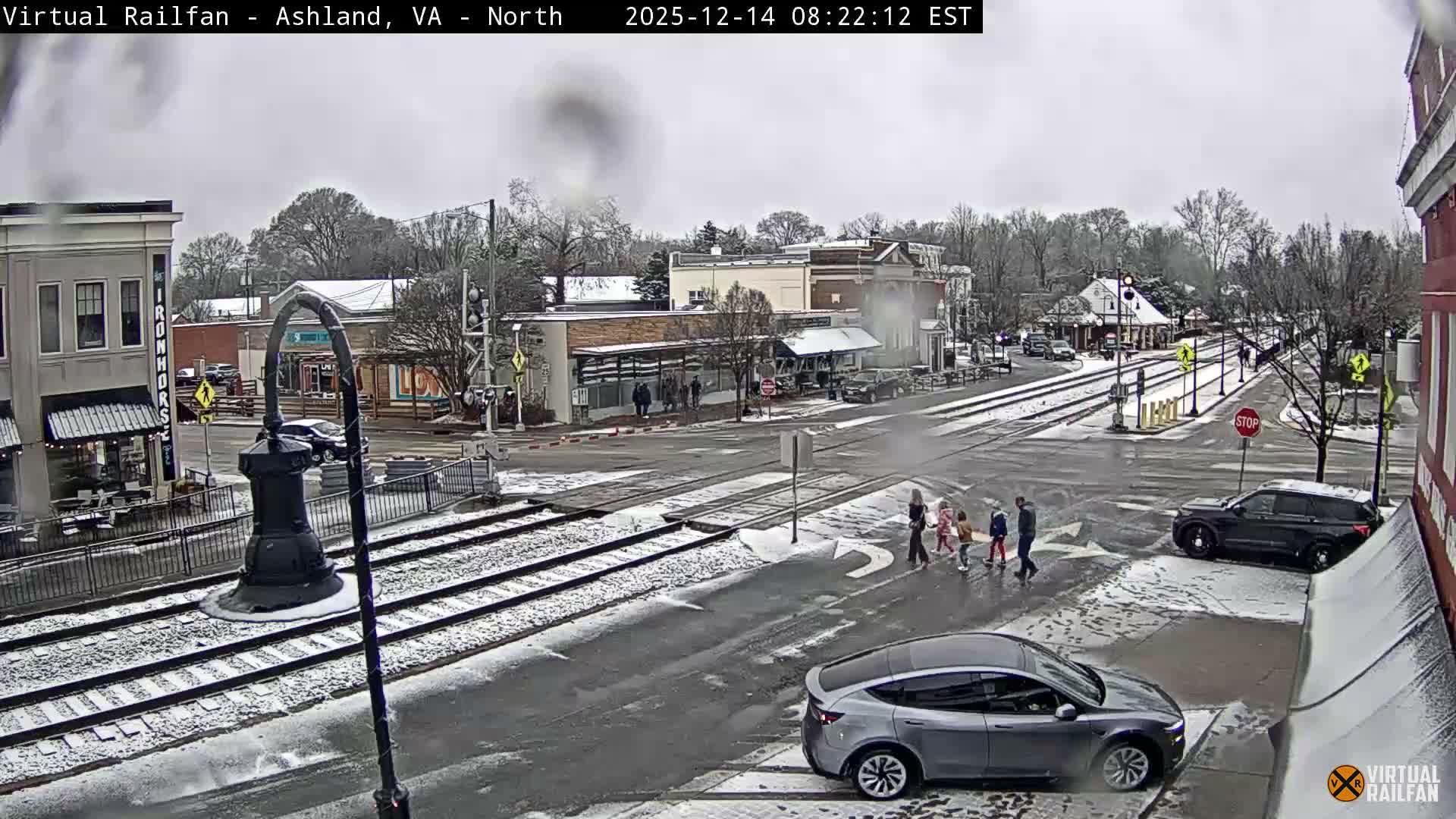 A wintry night scene shows a snow-covered town street with railroad tracks, buildings adorned with festive lights, and a vehicle, all illuminated under a blanket of fresh snow.