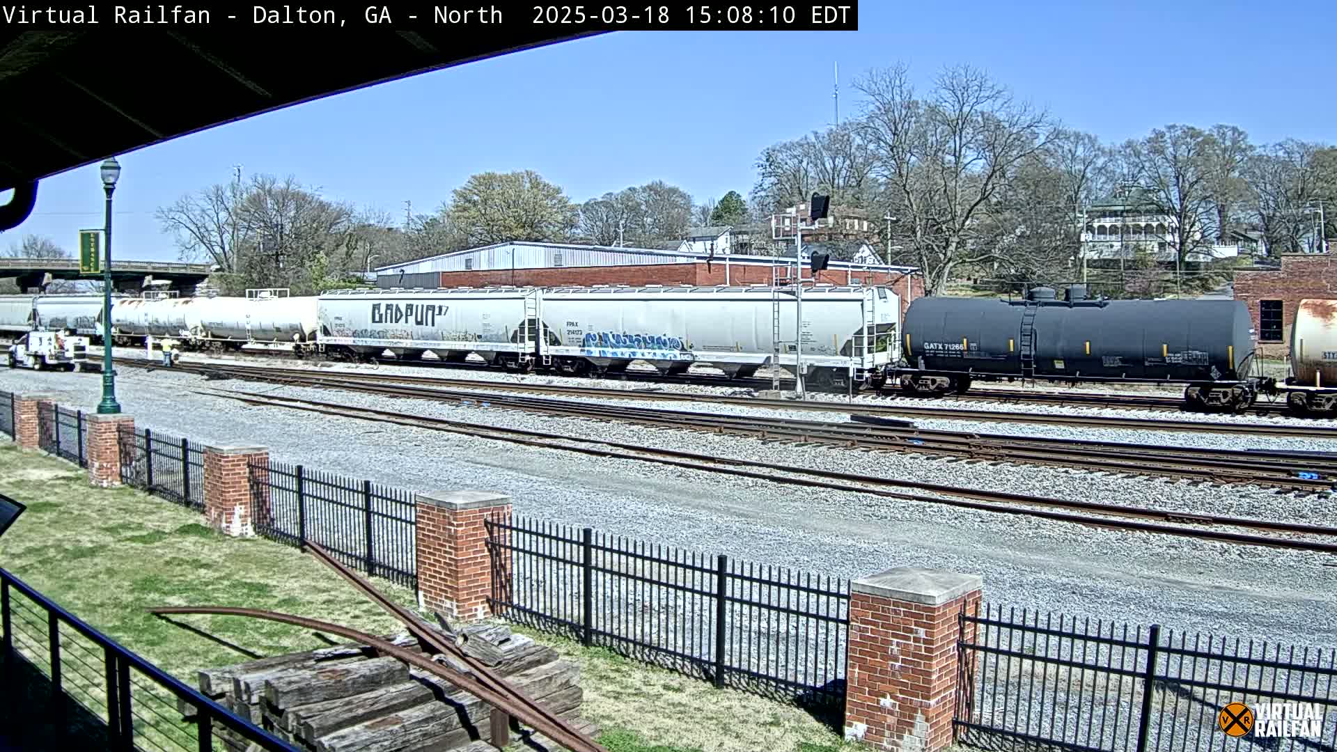 A freight train, consisting of several tanker cars and covered hopper cars, sits on a railway track under a clear, sunny sky.