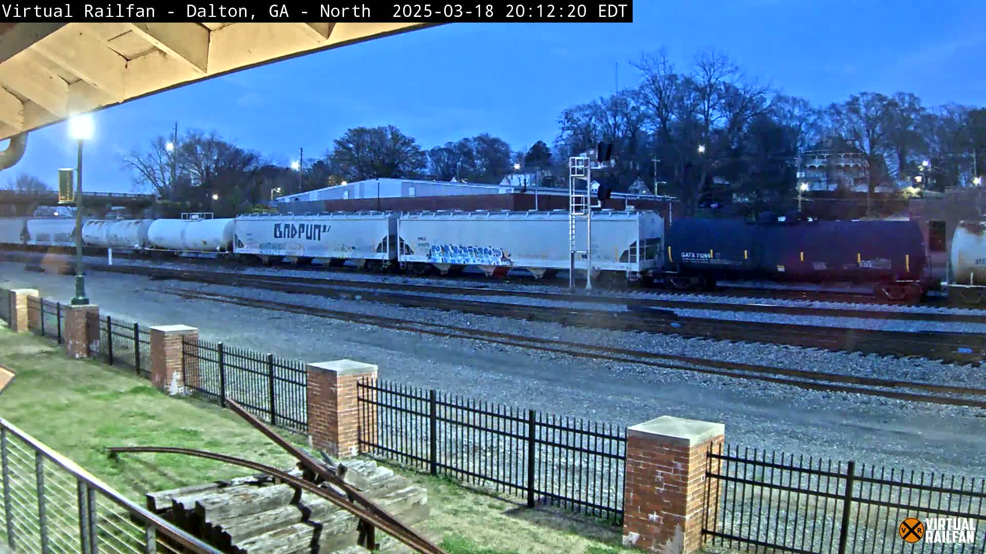 A freight train sits on a track near a brick fence under a twilight sky.