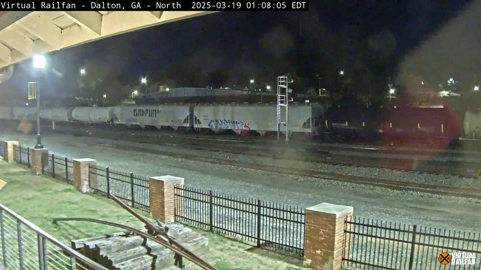 At night, a freight train with several cars passes by a metal fence and brick pillars near a grassy area.
