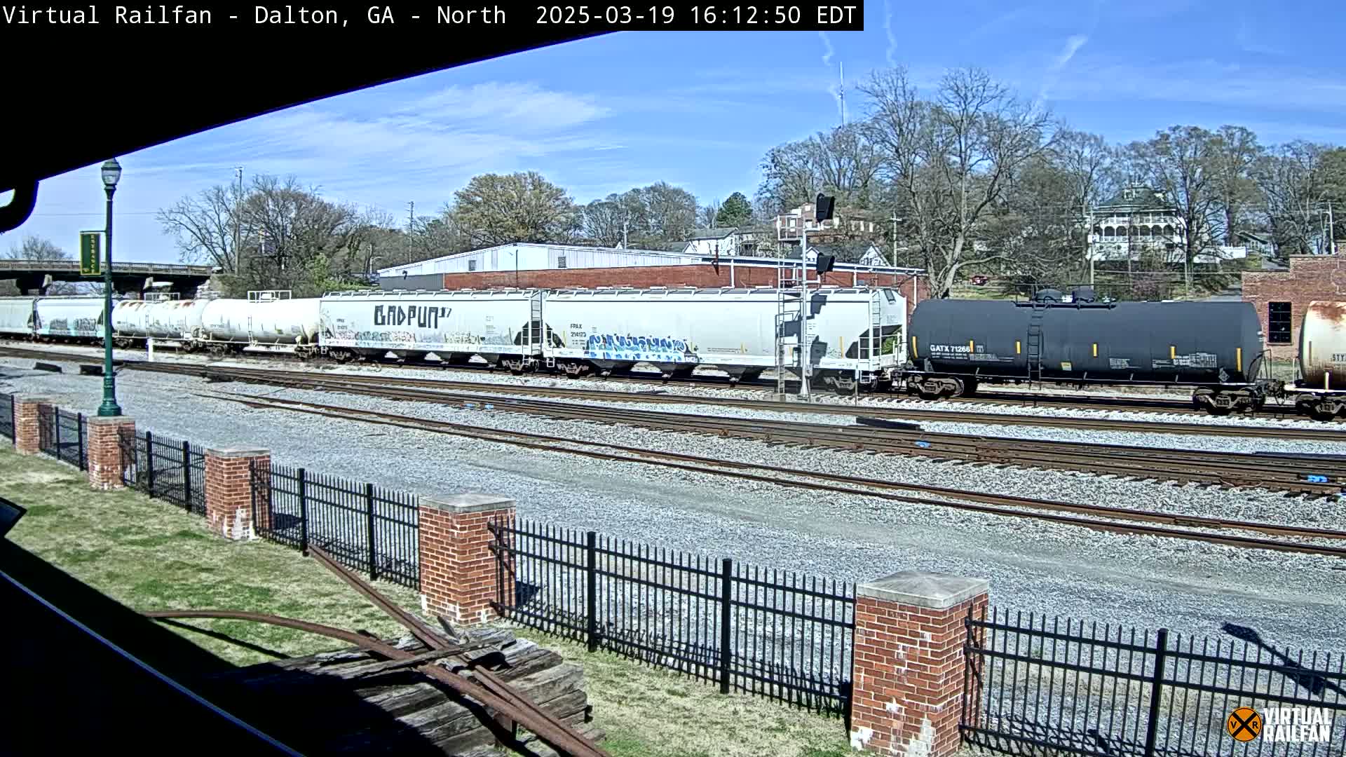 A long freight train sits on railroad tracks under a sunny sky, next to a brick fence and some grass.