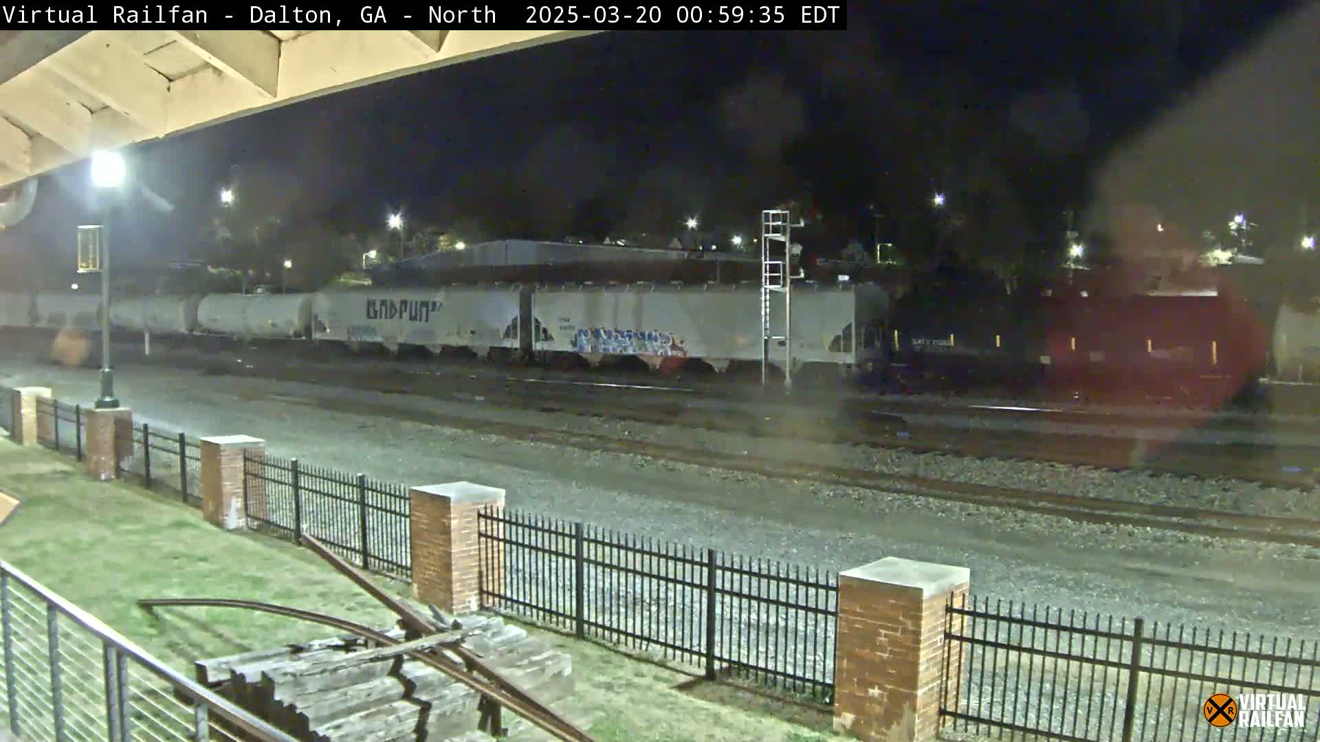 A freight train passes at night under a bridge, next to a brick fence and grassy area.