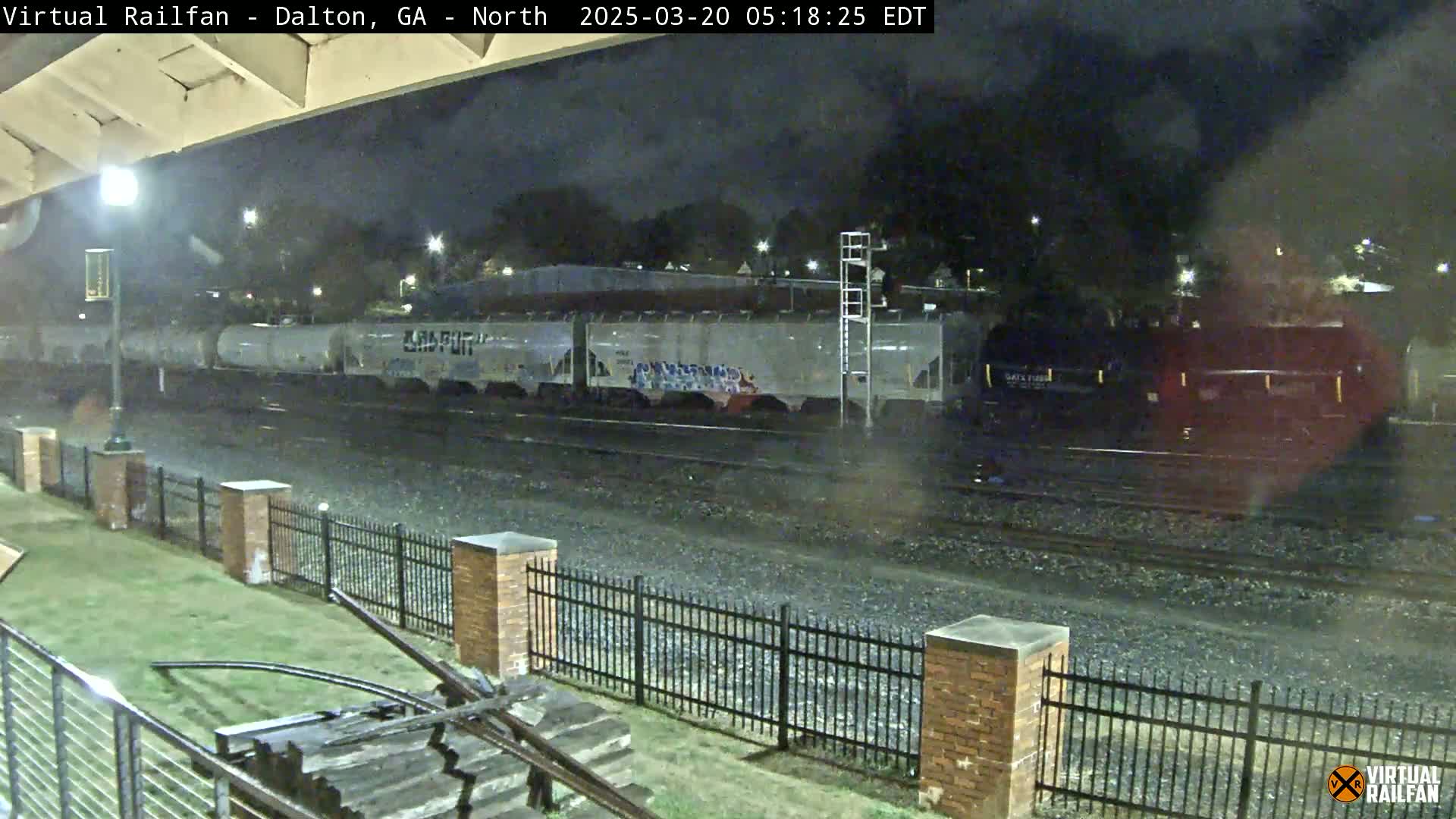 At night, a freight train passes by a section of railroad track next to a metal fence and grassy area.