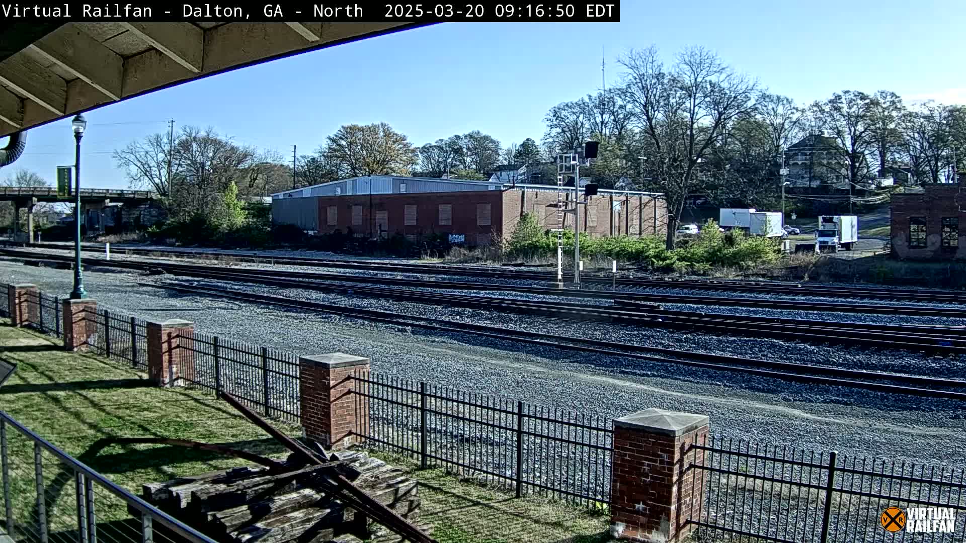 A sunny day reveals multiple train tracks extending into the distance, bordered by a brick fence and a grassy area, with buildings and trees visible in the background.