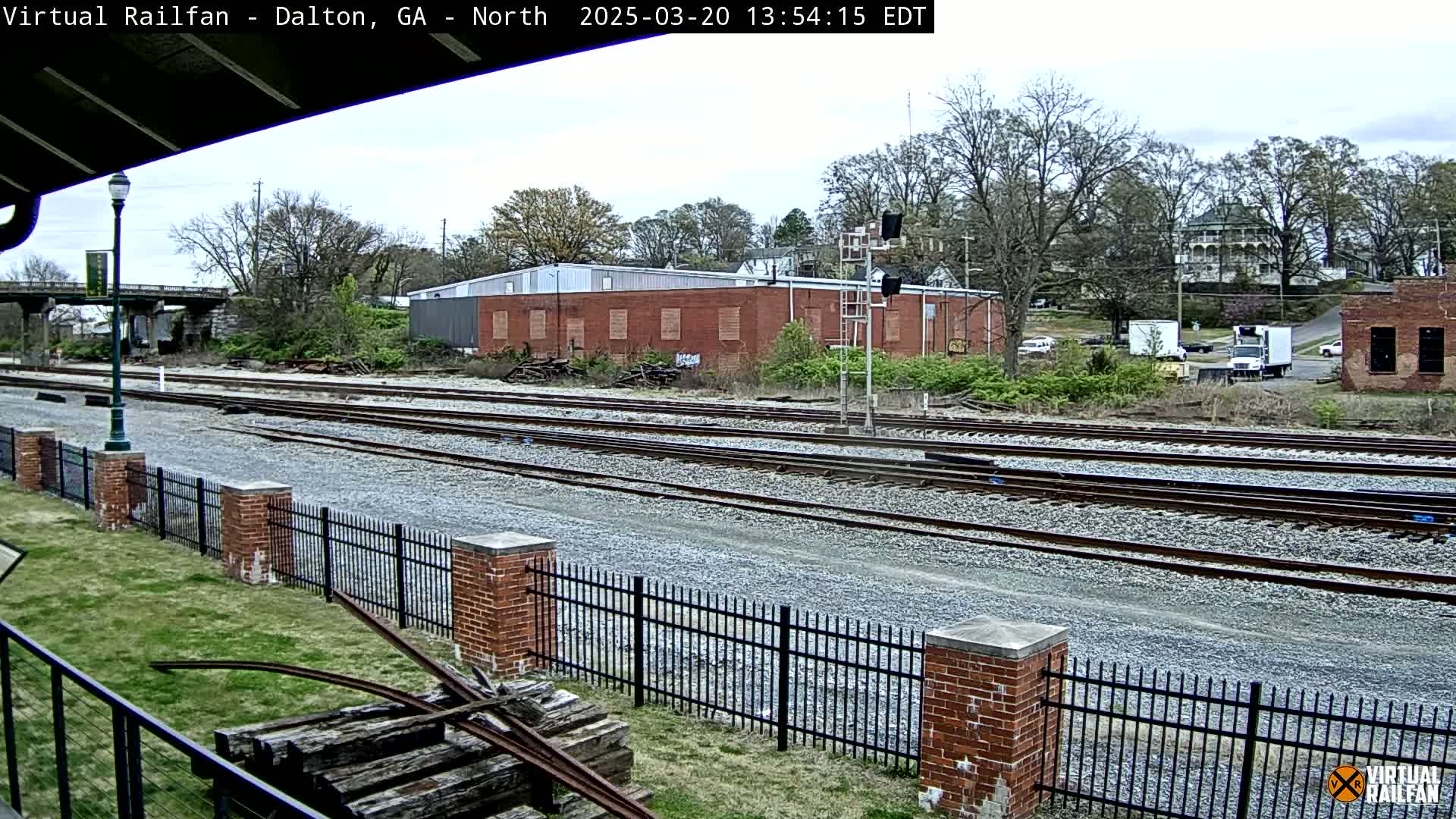 A view of several train tracks bordered by a brick fence on a partly cloudy day, with buildings and trees in the background.