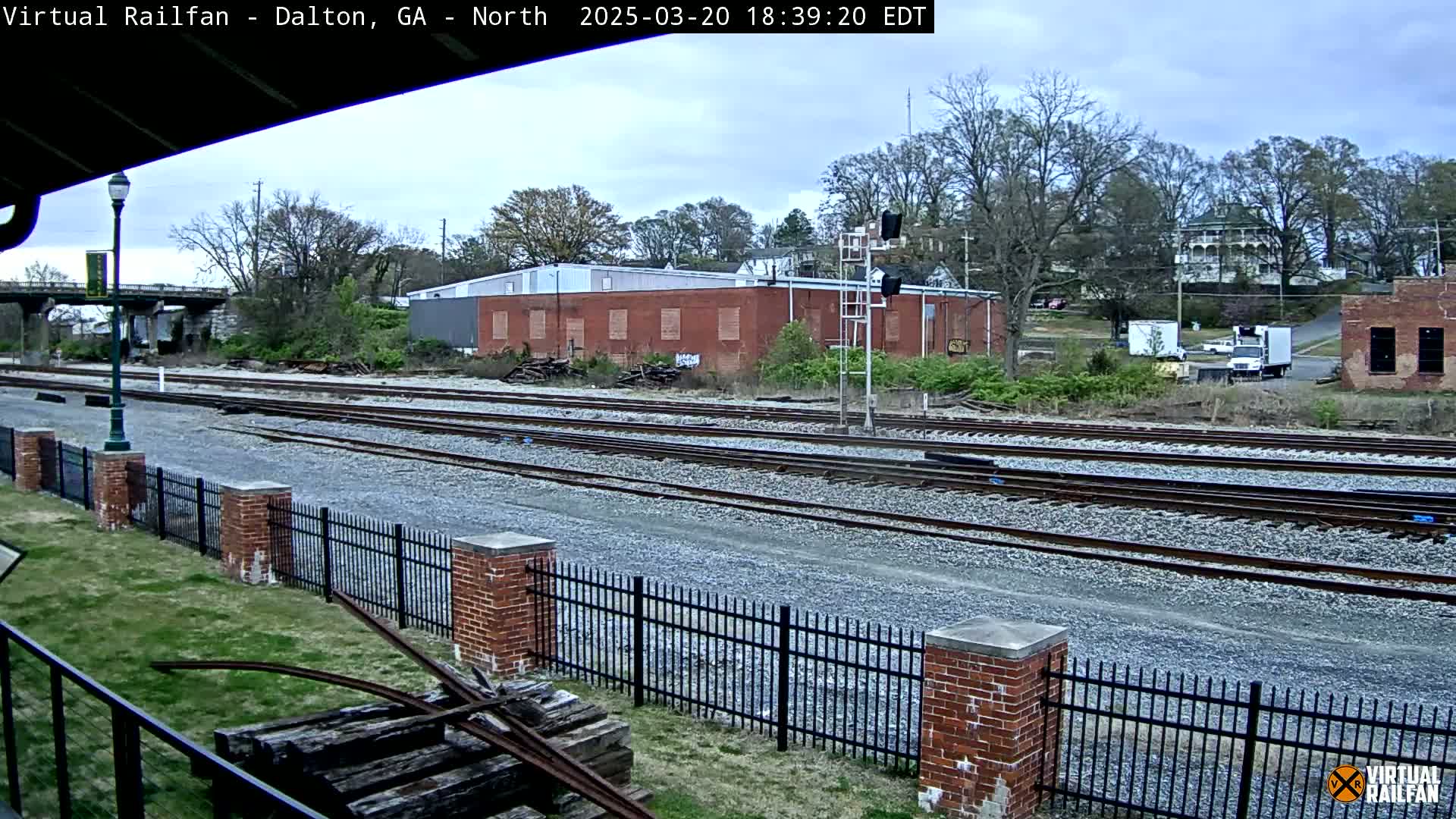 Multiple sets of train tracks extend into the distance under an overcast sky, bordered by a brick fence and surrounded by trees and industrial buildings.