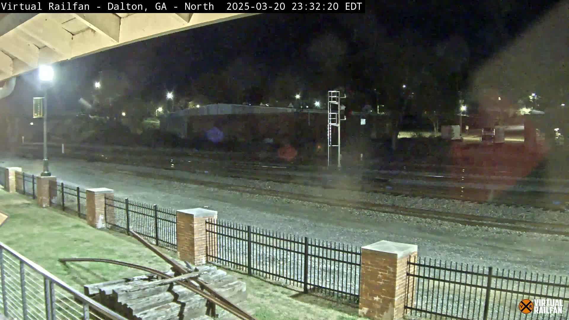A nighttime view of railroad tracks behind a short brick fence, with a section of old railway equipment visible in the foreground.