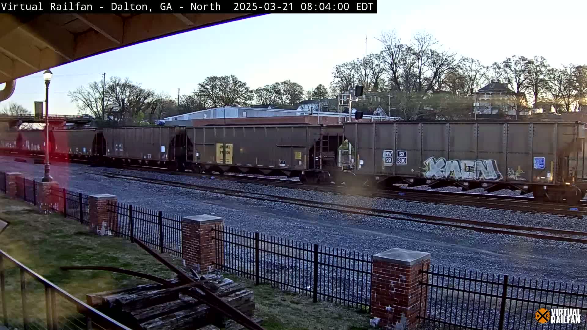 A long freight train of dark-colored cars passes by a brick fence under a partly cloudy morning sky.