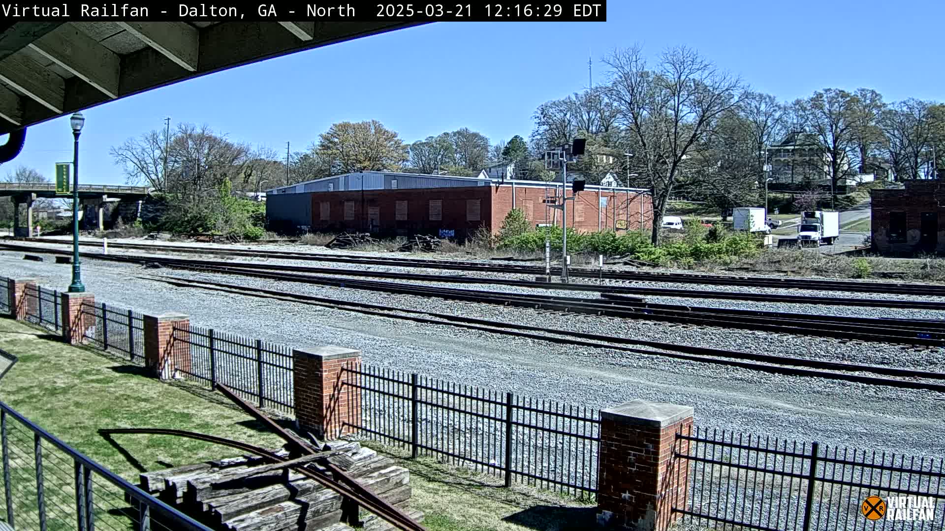 A sunny day reveals multiple train tracks extending into the distance, bordered by a brick fence and grassy area, with a brick building and trees in the background.