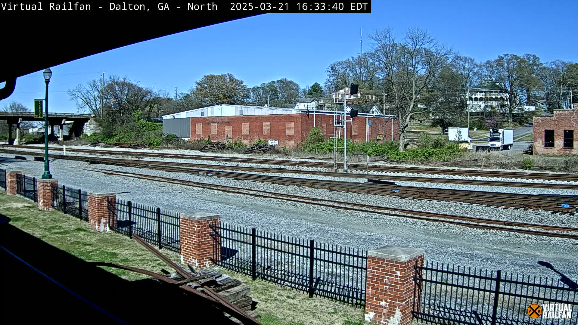 A sunny day reveals multiple train tracks bordered by a brick fence, with a brick building and trees visible in the background.