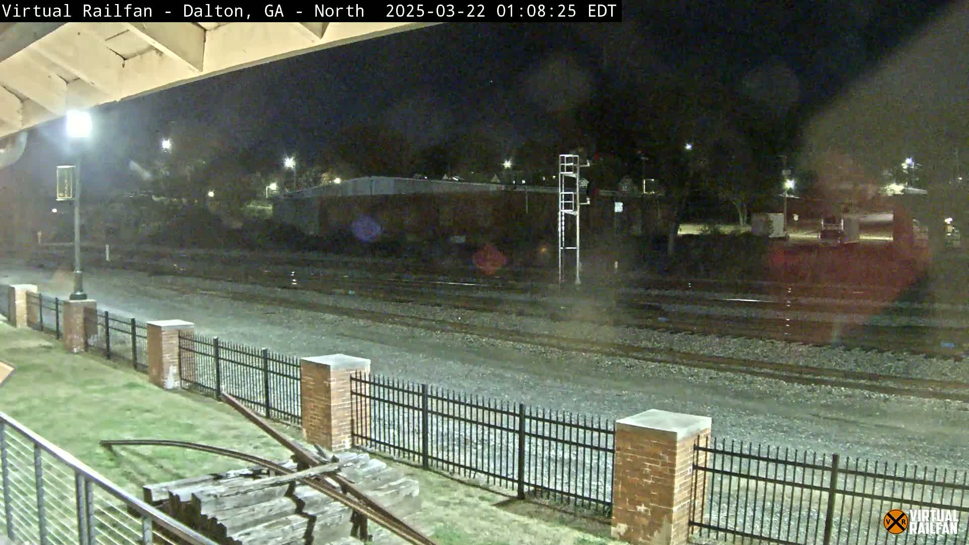 A nighttime view of railroad tracks, bordered by a brick fence and a grassy area, under a clear, dark sky.
