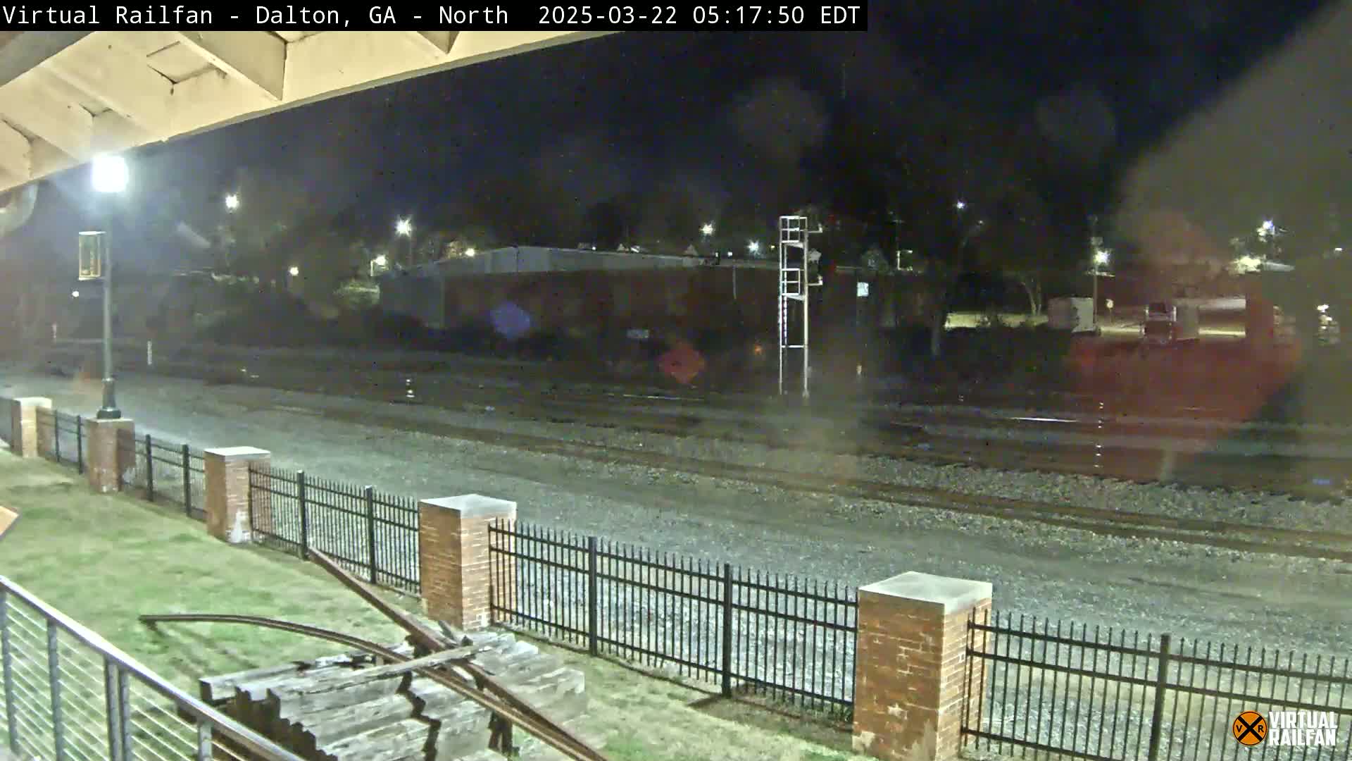 A nighttime view of railroad tracks, a brick fence, and some vegetation, under dark and possibly slightly overcast conditions.
