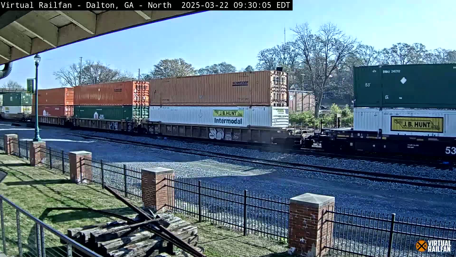 A freight train carrying multiple shipping containers, some stacked two high, passes by on a sunny day.