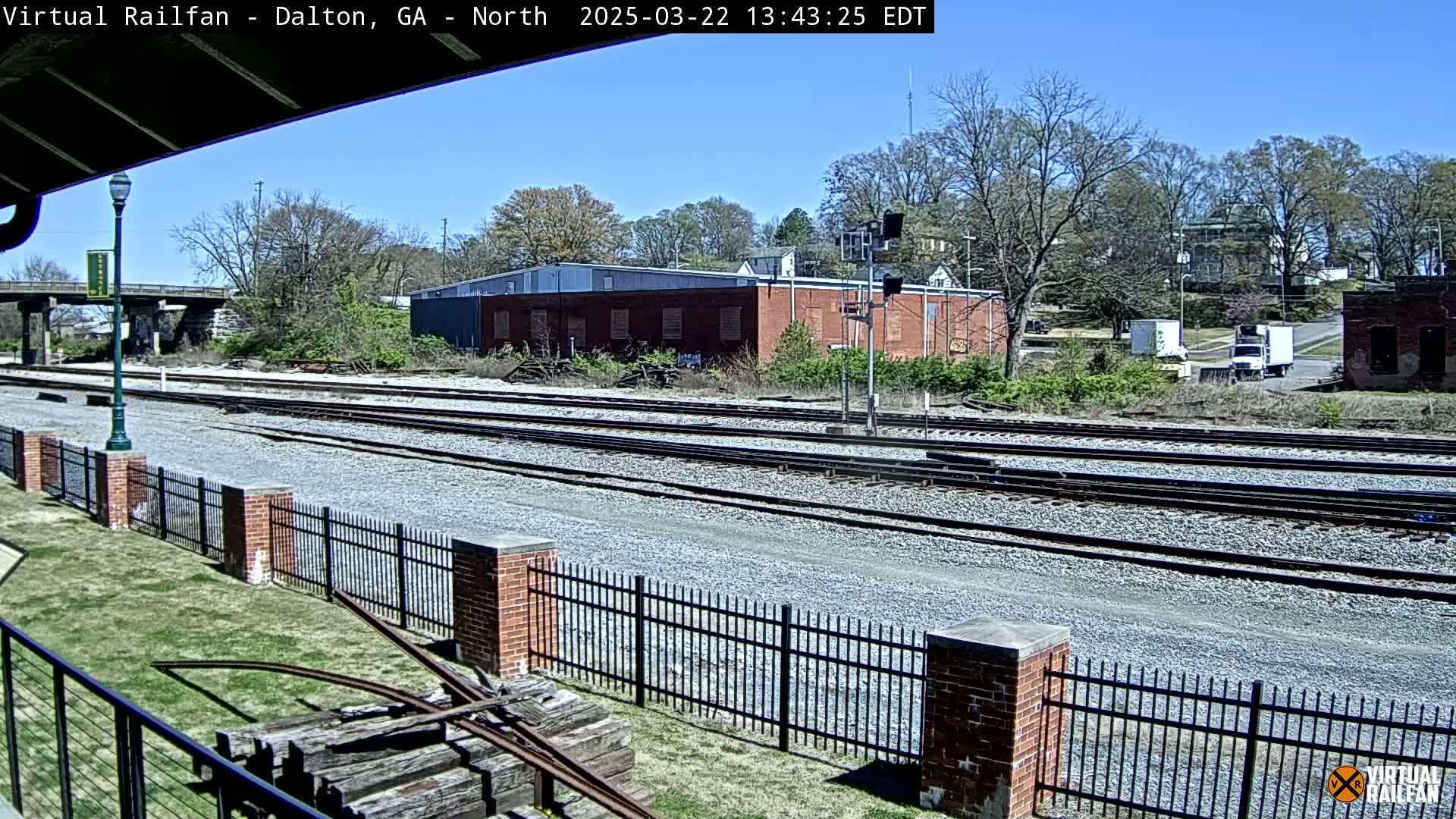 A sunny day reveals multiple train tracks bordered by a brick fence and overgrown vegetation, with a brick building and trees in the background.