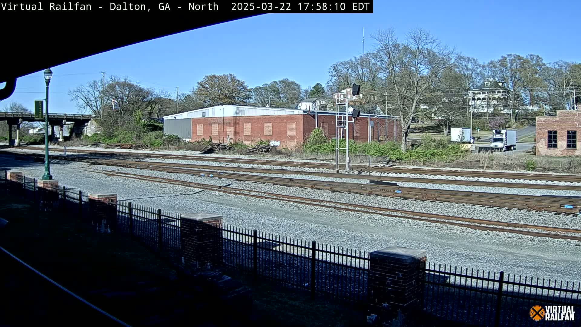 A sunny view of several railway tracks next to a brick building and a chain-link fence, with trees and other buildings in the background.
