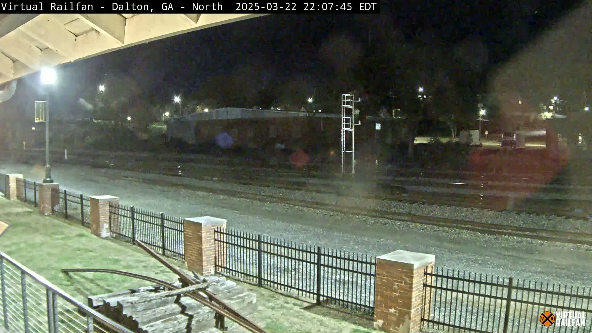A nighttime view of railroad tracks, bordered by a brick fence and a grassy area, under a clear, dark sky.